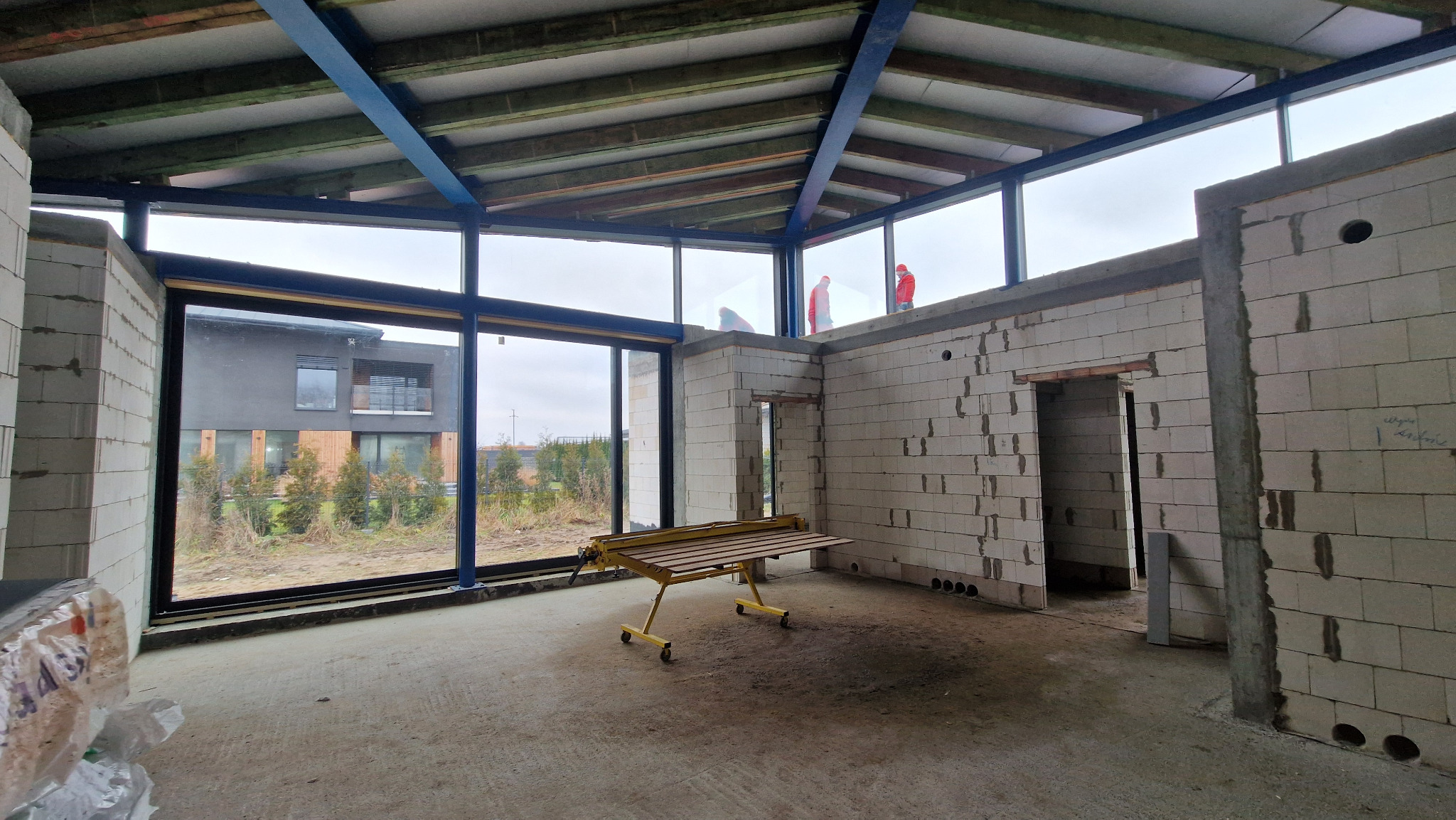 Surowy stan wnętrza z dużymi oknami z ramą w kolorze granatowym, widok na zewnątrz. Na górze budynku dwóch robotników w czerwonych kombinezonach.