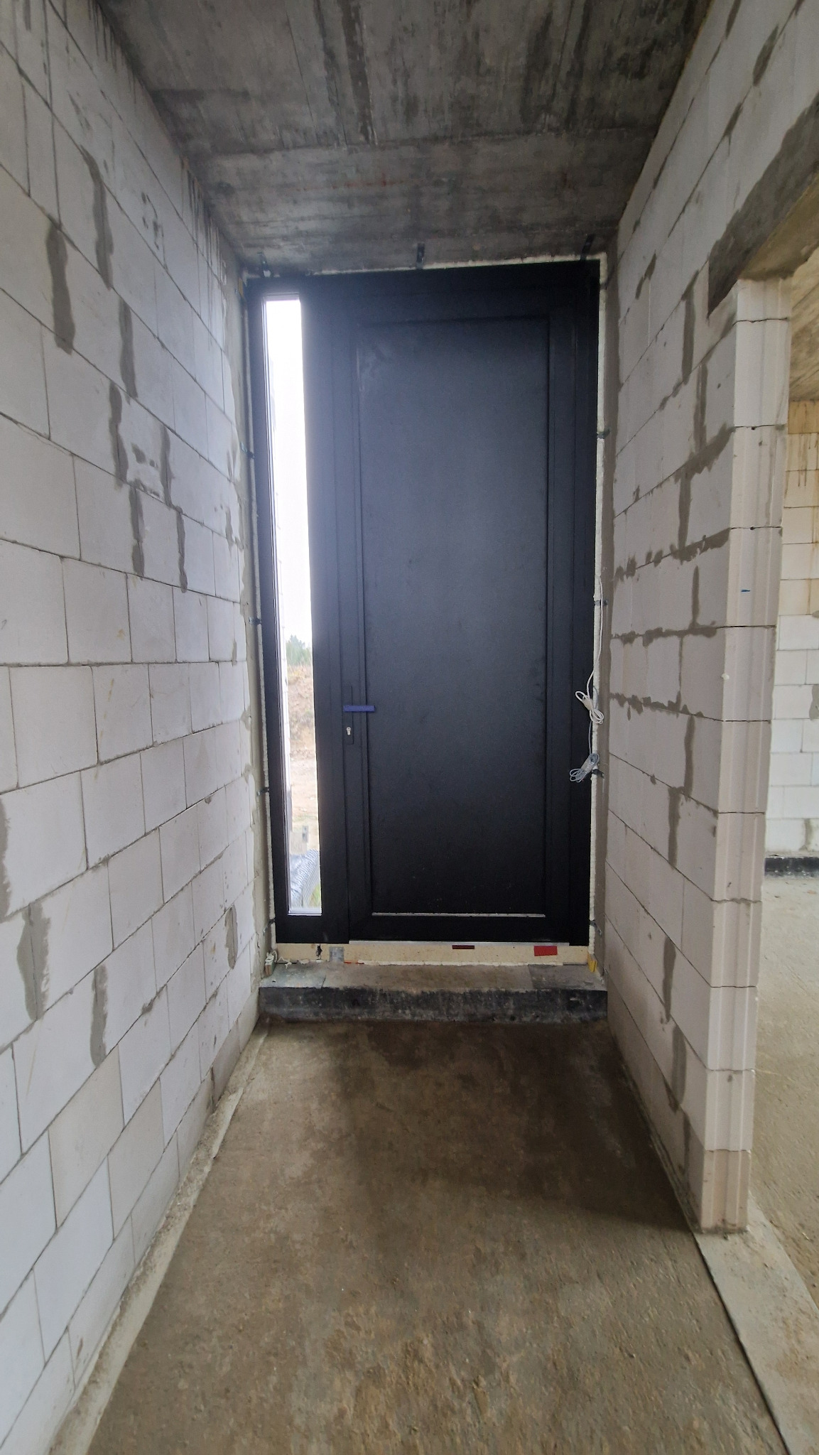 Ciemne drzwi z wąskim oknem bocznym, zamontowane w surowym wnętrzu z białych bloczków. Widoczne kable i surowy betonowy strop.