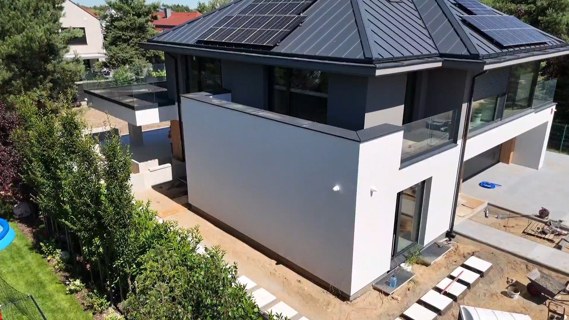 Nowoczesny dom z panelami słonecznymi na dachu, dużymi oknami i szklanymi balustradami balkonów. Widok z góry, otoczenie w trakcie prac wykończeniowych.