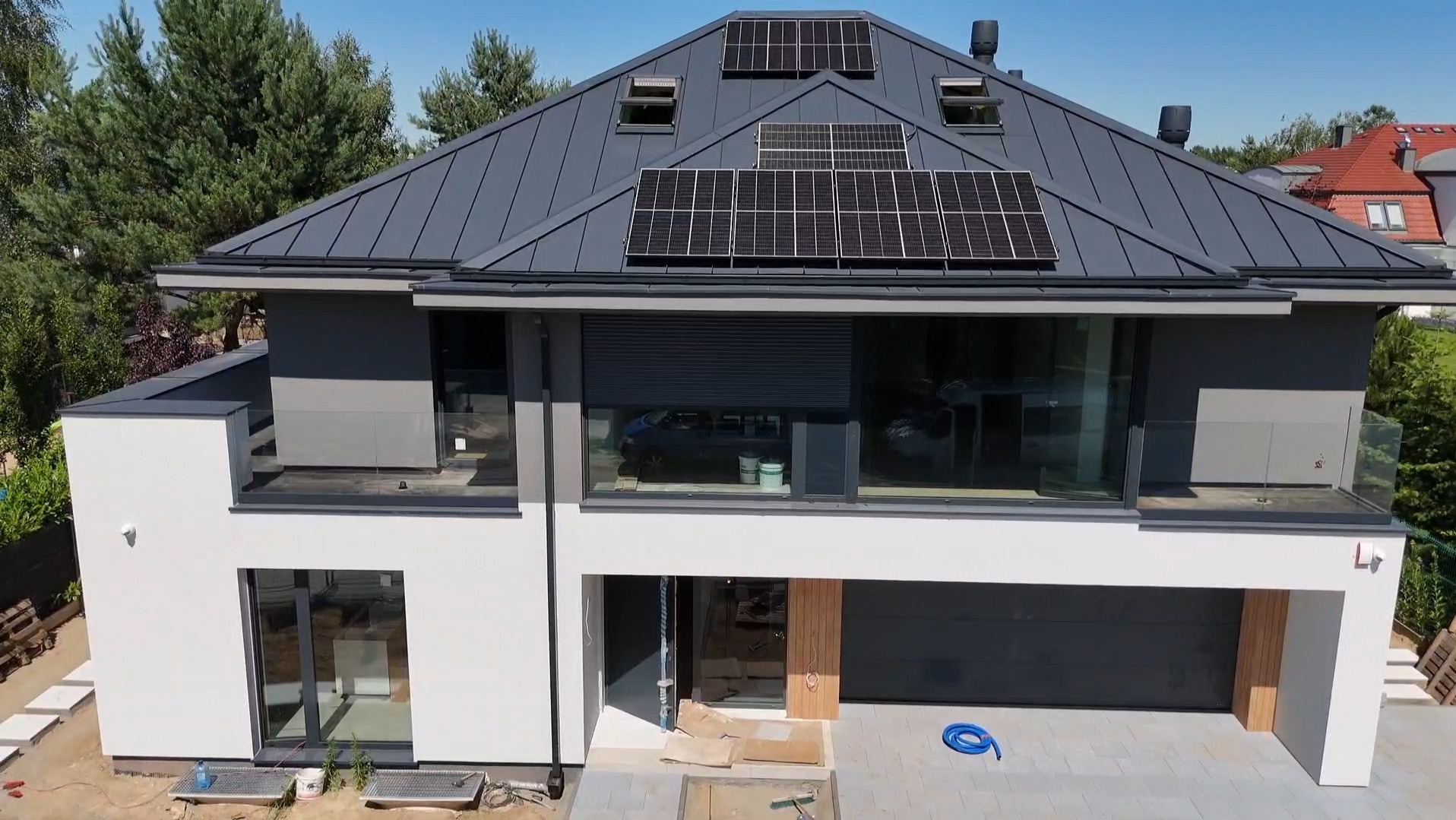 Nowoczesny dom z grafitowym dachem i panelami słonecznymi. Duże okna z roletami zewnętrznymi. Częściowo wykończony teren wokół budynku.