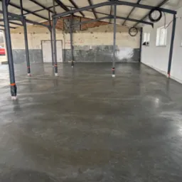 Świeżo wylana, gładka posadzka przemysłowa w hali z metalową konstrukcją i częściowo wykończonymi ścianami. Widoczne słupy z zabezpieczeniami.