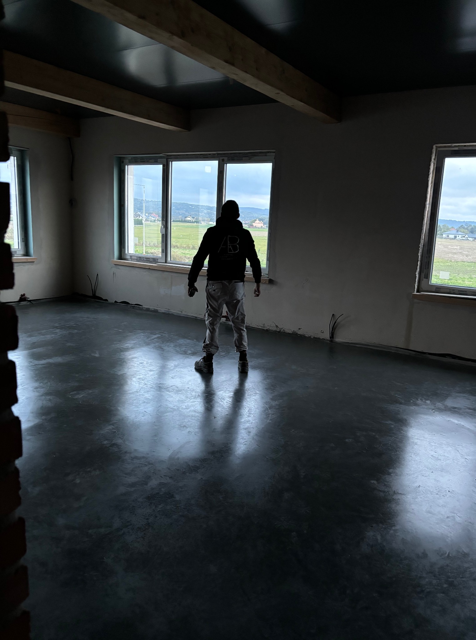 Wykończona, błyszcząca posadzka przemysłowa w surowym wnętrzu. Widoczne okna z widokiem na zewnątrz i osoba w oddali, stojąca na posadzce. Minimalistyczny styl.