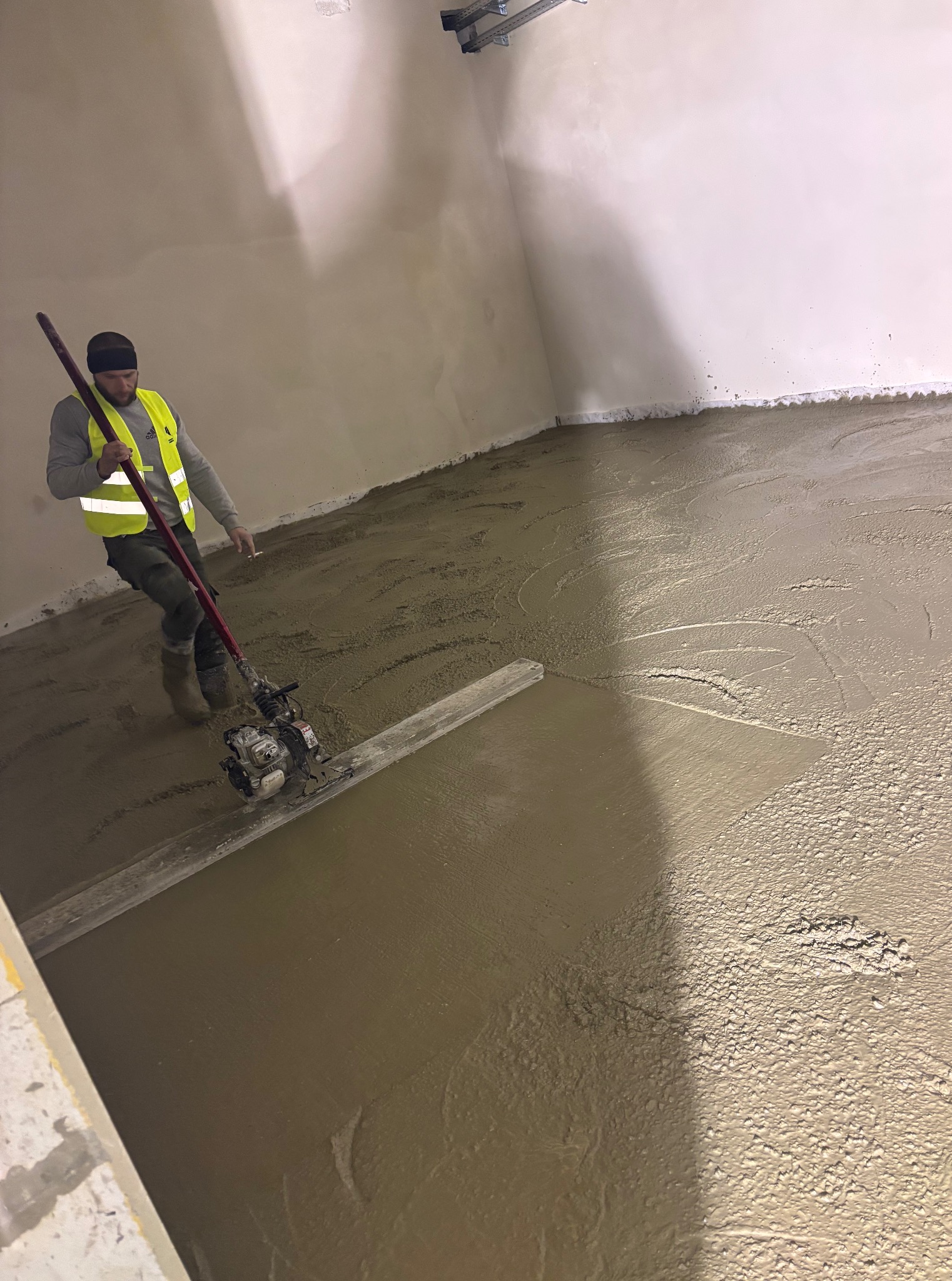 Pracownik w kamizelce wyrównuje wylewkę maszyną w pomieszczeniu. Świeża, szara posadzka przemysłowa w trakcie realizacji.