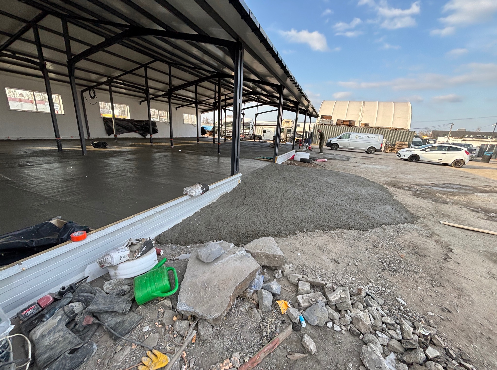 Wylewka świeżej, szarej posadzki w hali z metalową konstrukcją dachu, widoczne narzędzia i gruz budowlany obok. W tle zaparkowane samochody.
