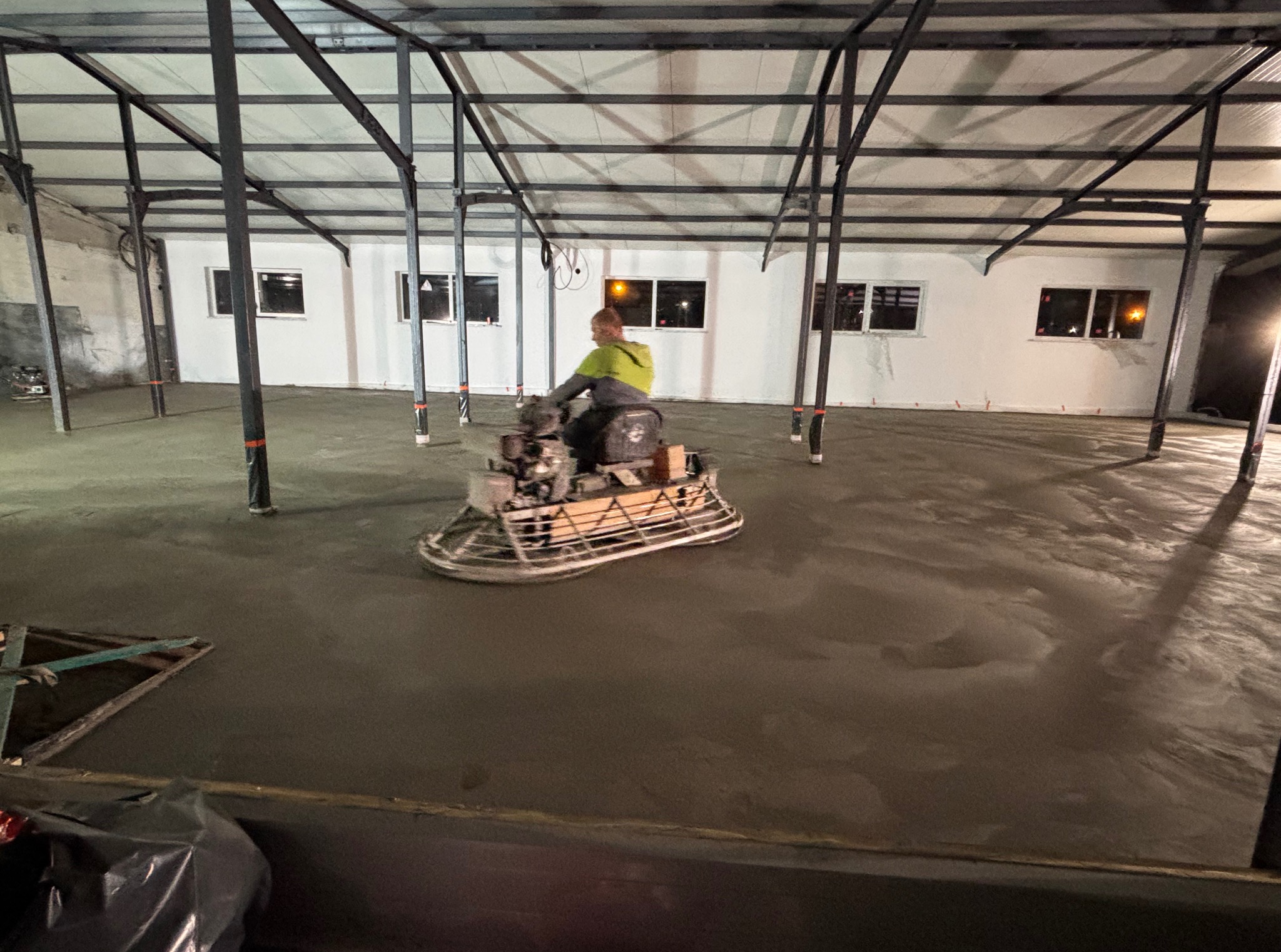 Pracownik w żółtej bluzie wyrównuje mokrą, szarą posadzkę przemysłową za pomocą zacieraczki mechanicznej w hali z metalową konstrukcją i białymi ścianami.