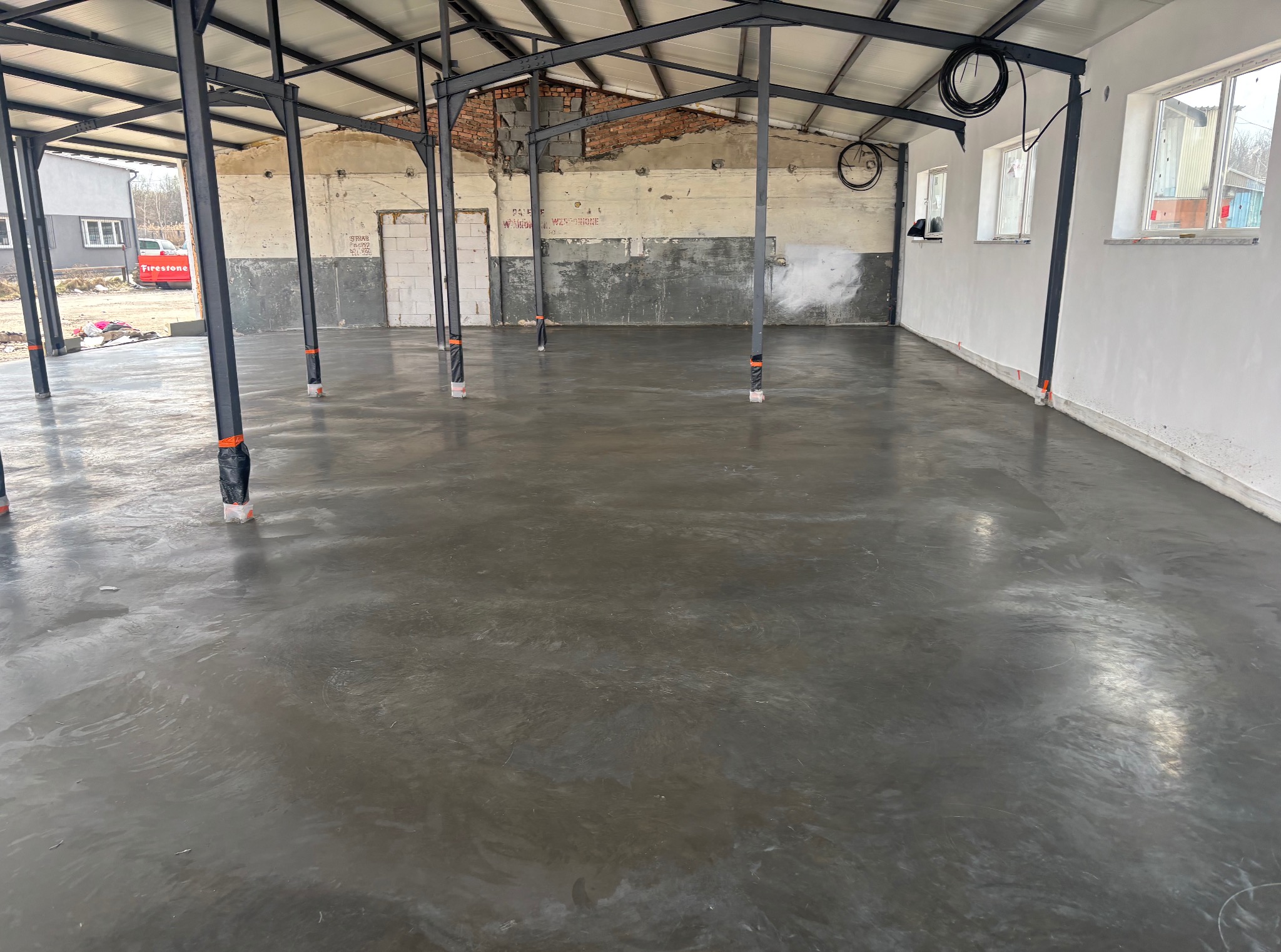 Świeżo wylana, gładka posadzka przemysłowa w hali z metalową konstrukcją i częściowo wykończonymi ścianami. Widoczne słupy z zabezpieczeniami.