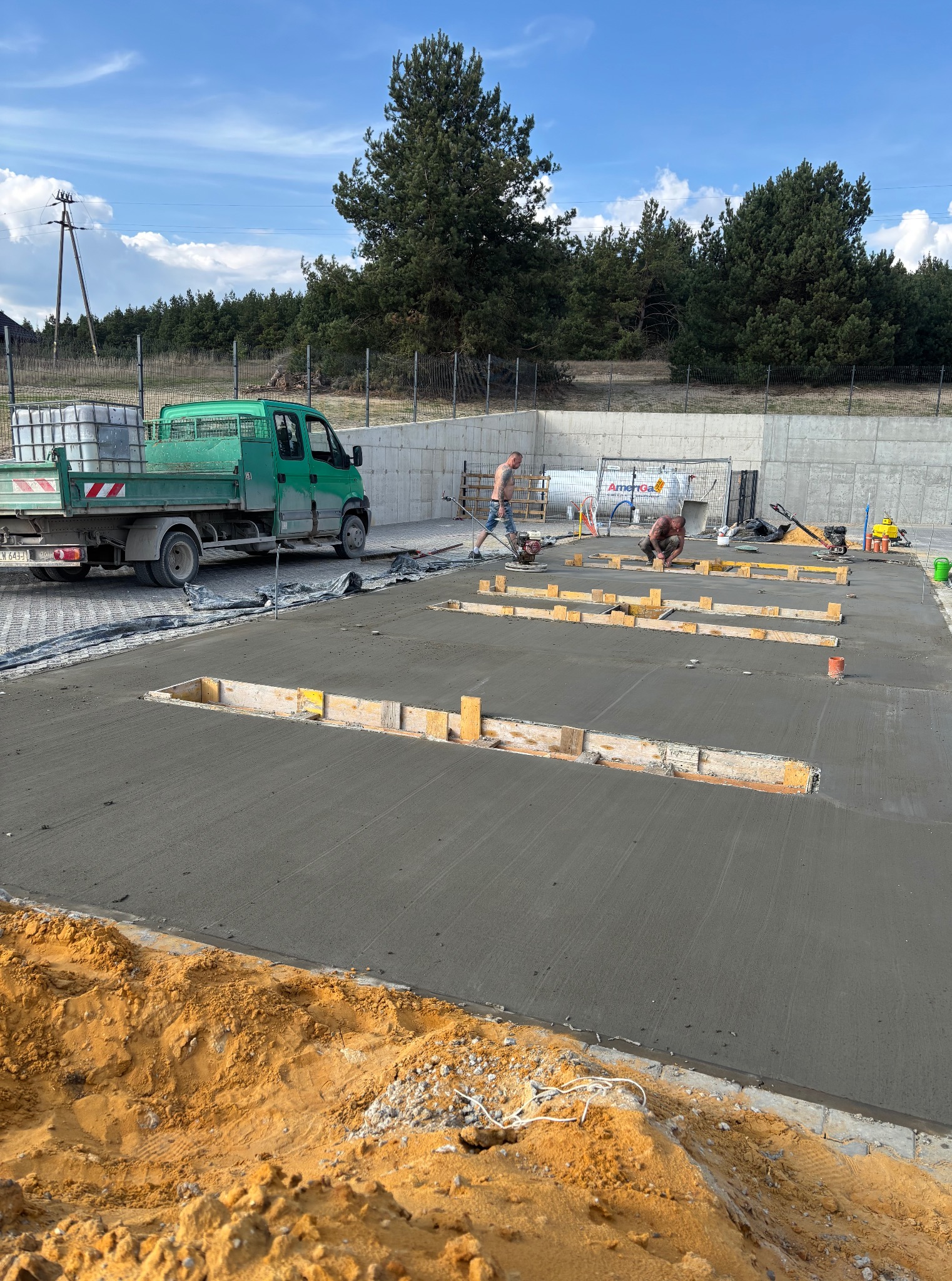 Wylewanie betonu na placu budowy z drewnianymi szalunkami. W tle zielony samochód i dwóch pracowników podczas pracy nad posadzką. Słoneczny dzień.