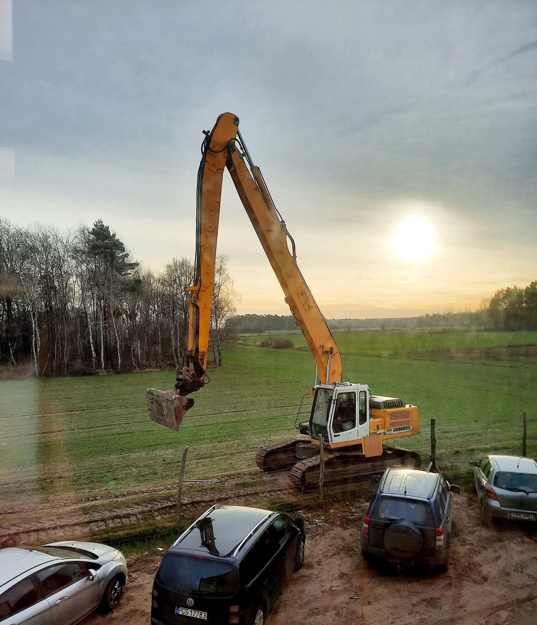 Żółto-biała koparka LIEBHERR na polu z widocznymi śladami pracy, w tle las i zachodzące słońce, na pierwszym planie zaparkowane samochody.
