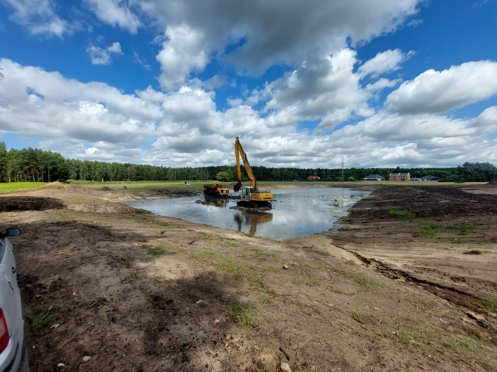 Żółta koparka Liebherr na wodzie w wykopanym stawie, odbicie chmur w wodzie. W tle las i zabudowania.