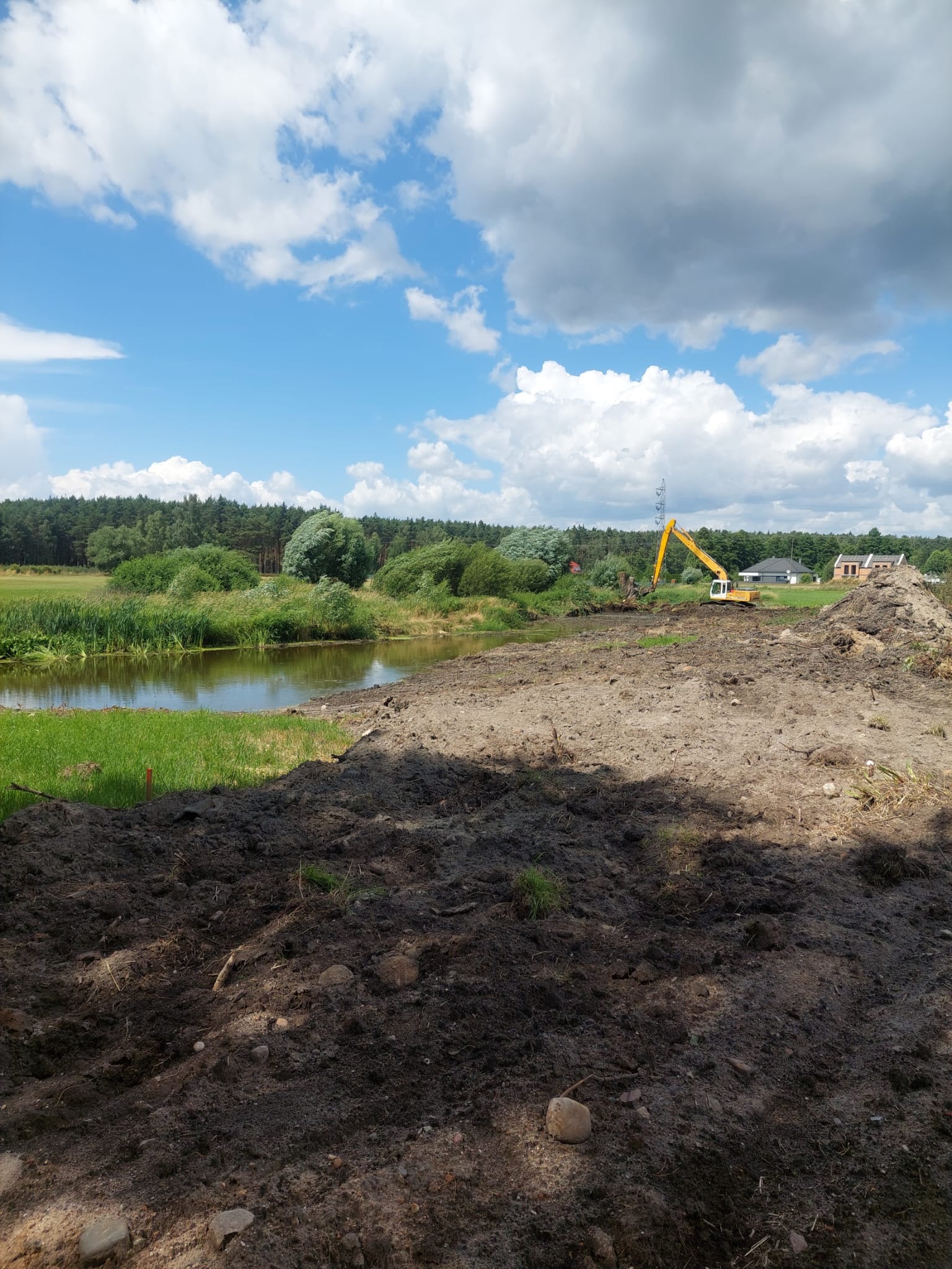 Żółta koparka nad brzegiem rzeki w trakcie prac ziemnych. W tle drzewa i budynek. Błękitne niebo z chmurami.
