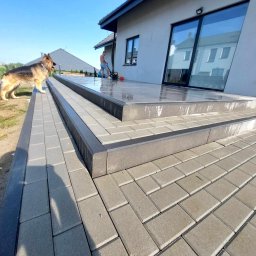 Łukasz Danielewski - Ułożona kostka brukowa w odcieniach szarości tworzy schody tarasowe. W tle mężczyzna czyści powierzchnię myjką ciśnieniową, obok stoi owczarek niemiecki.