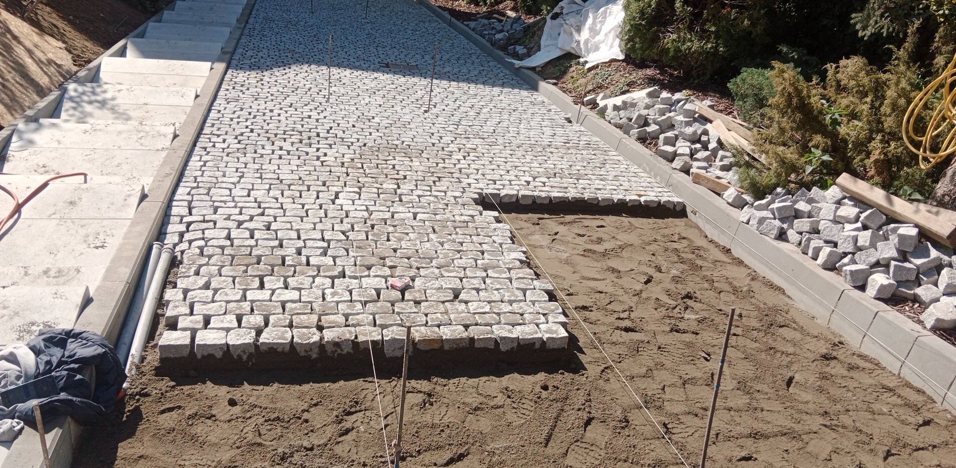 Fragment ścieżki z kostki granitowej w trakcie układania na nierównym terenie. Część powierzchni wyłożona, widoczne narzędzia i zapas kostki. Schody w tle.