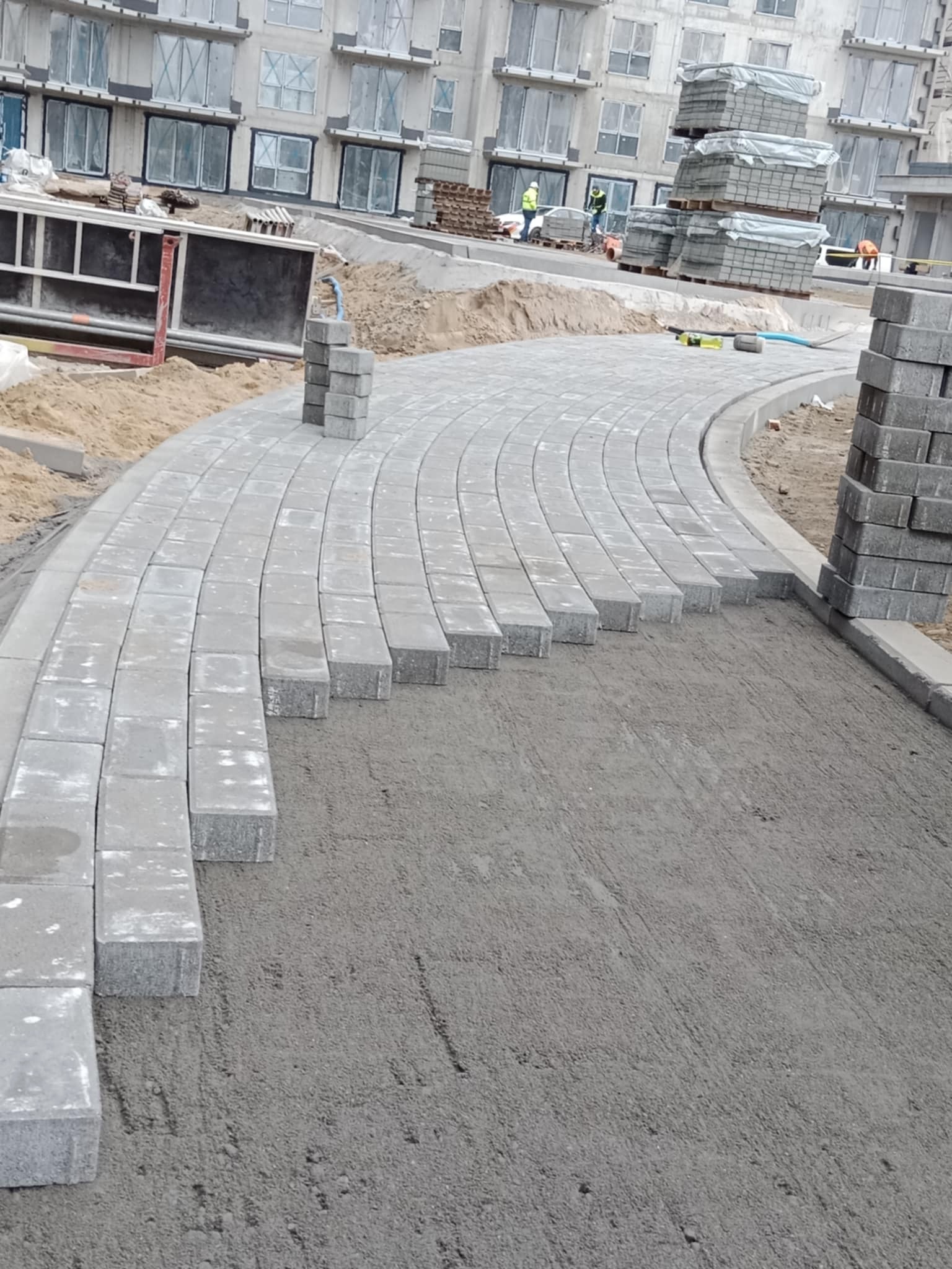 Etap układania szarej kostki brukowej w łuk na przygotowanym podłożu. W tle budynek w trakcie budowy i stosy kostki brukowej.