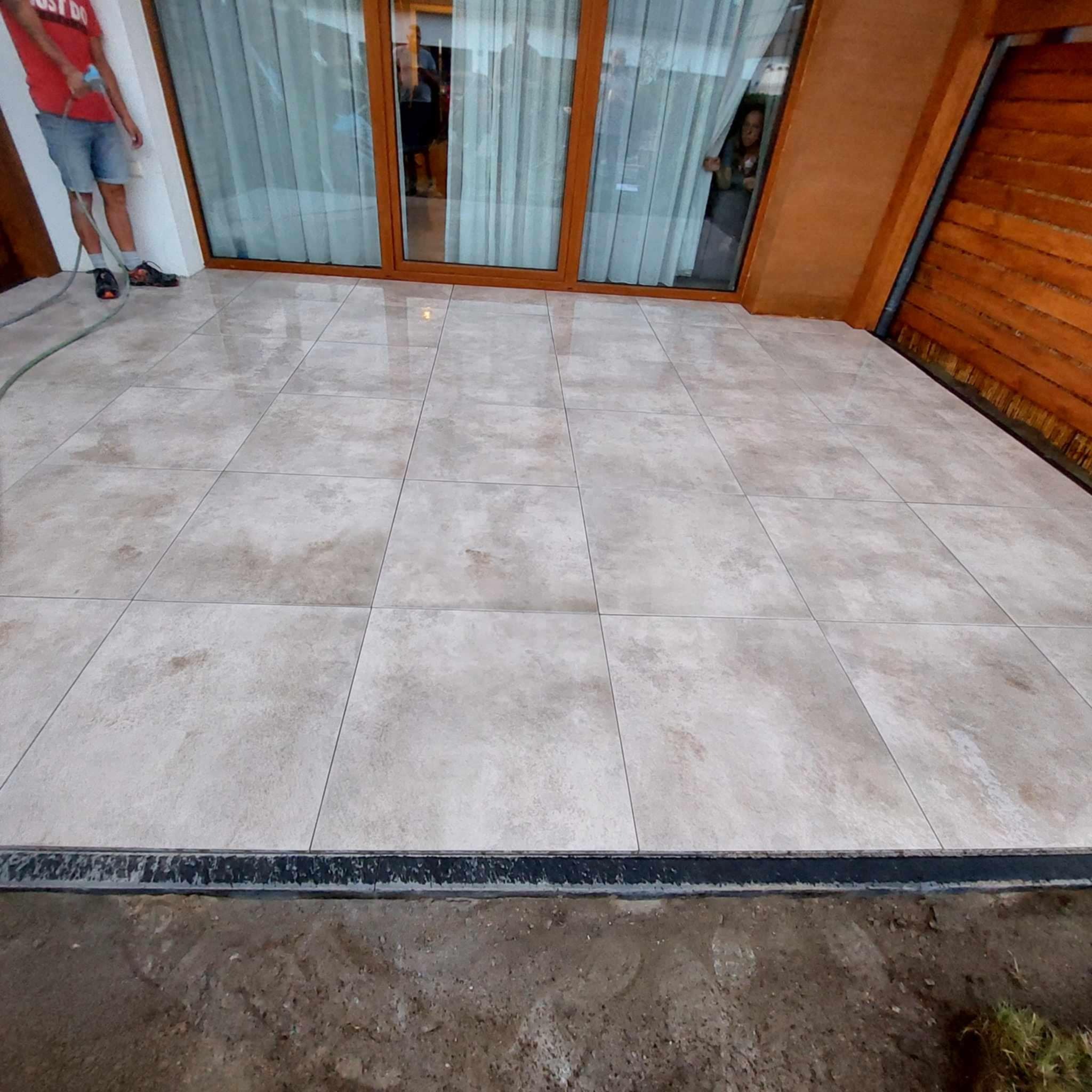 Jasne płytki podłogowe na tarasie, widok z dołu. W tle okno tarasowe i fragment drewnianej elewacji. Na pierwszym planie widoczna krawędź tarasu i ziemia.