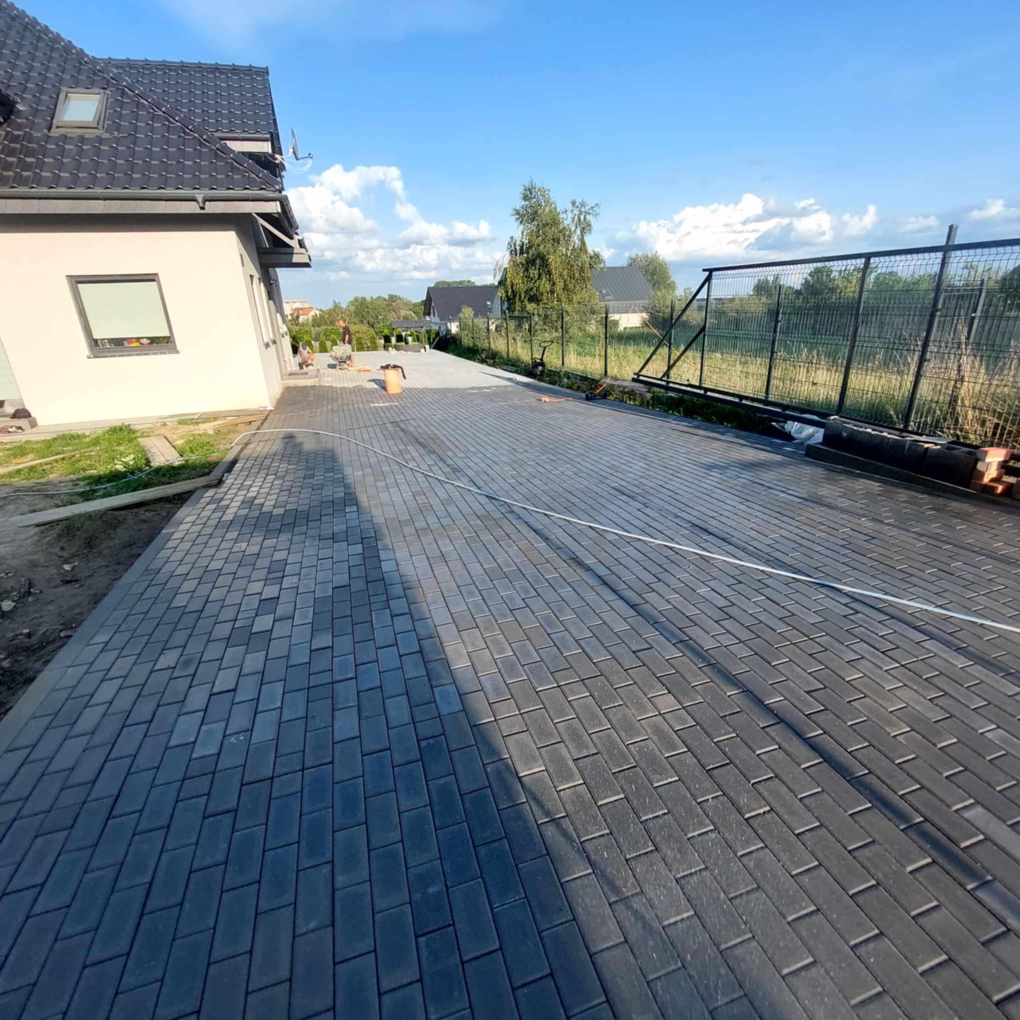 Ułożona, grafitowa kostka brukowa na podjeździe domu jednorodzinnego. W tle widoczni pracownicy i ogrodzenie. Słoneczny dzień.