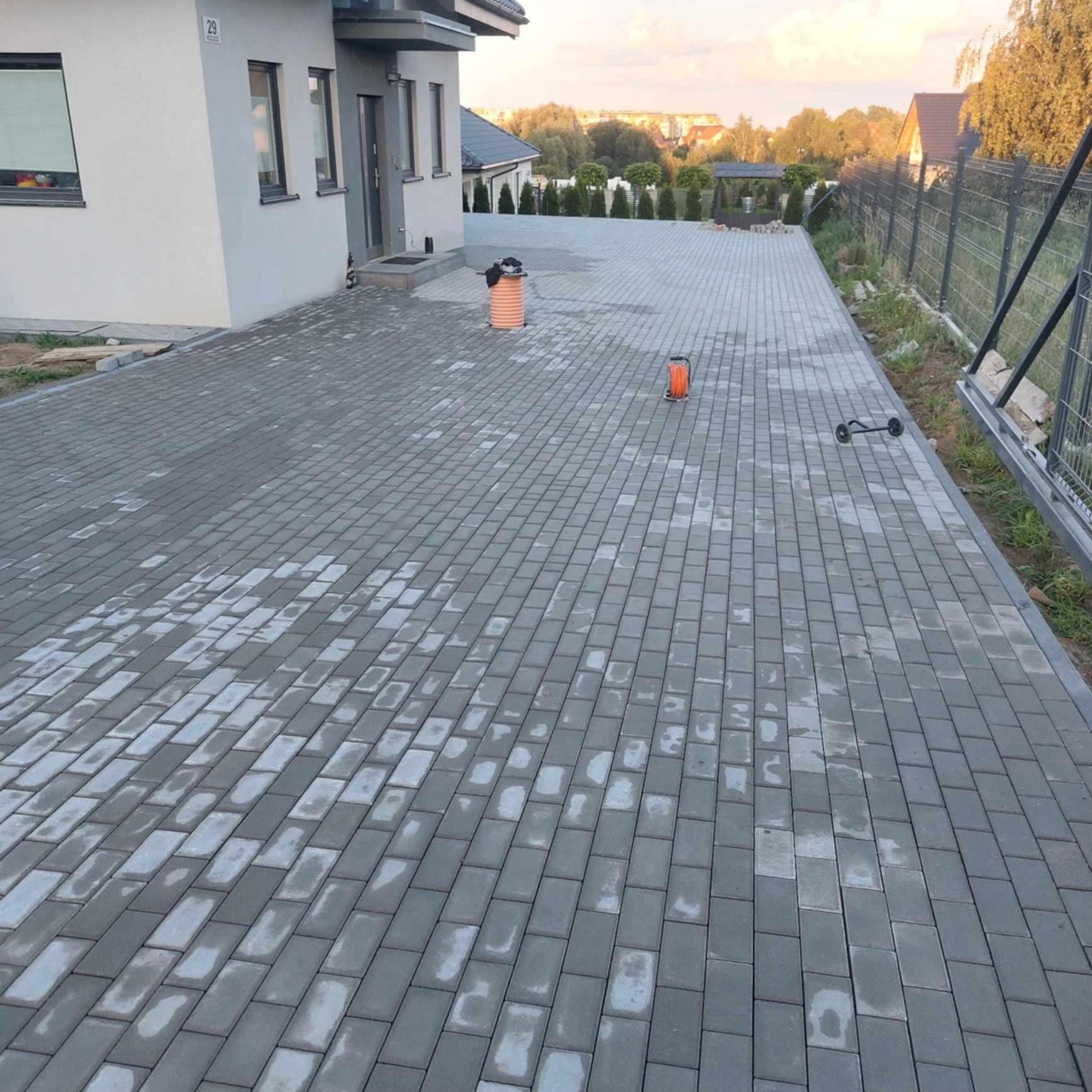 Szeroki podjazd z szarej kostki brukowej prowadzący do domu z numerem 29. Widoczne narzędzia: zwinięty kabel i rura. W tle zabudowa jednorodzinna i zieleń.