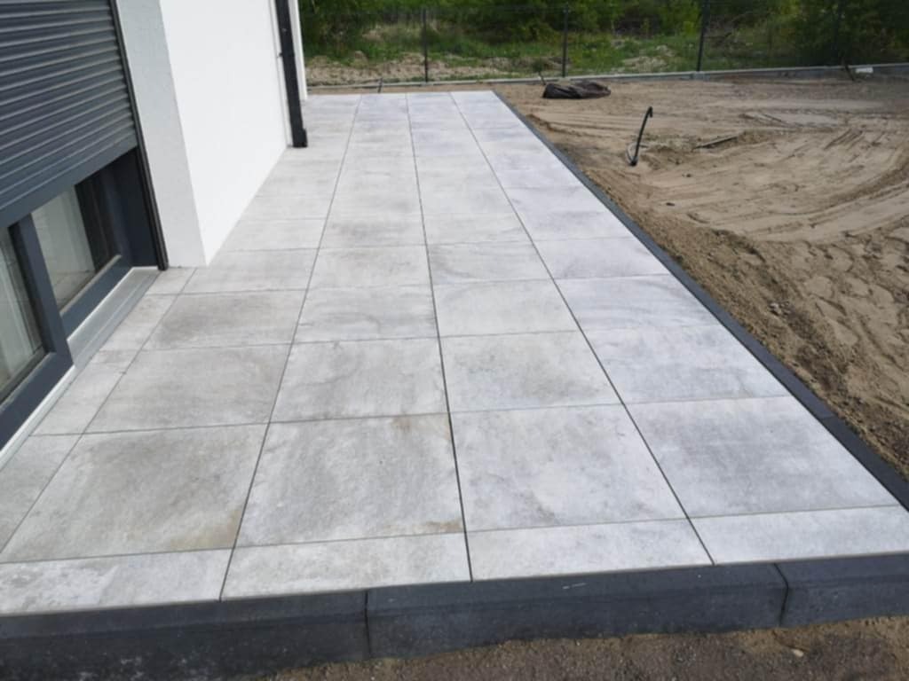 Jasnoszara kostka brukowa ułożona na tarasie przy budynku z szarą roletą okienną. Widoczny fragment trawnika. Geometryczny wzór kostki.