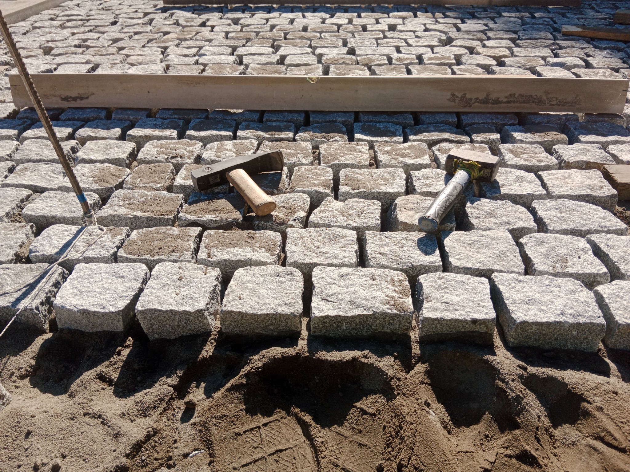 Fragment układania kostki granitowej: poziomica, młotki i sznur na piasku. Kostka ułożona częściowo, widoczne narzędzia pracy, surowy piasek jako podłoże.