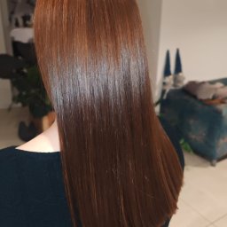 PRACOWNIA FRYZJERSKA ANITA DRATKOWICZ - Lśniące, proste włosy w odcieniach miedzi, widoczne od tyłu, po zabiegu koloryzacji. W tle fragment wnętrza z rośliną i turkusową sofą.