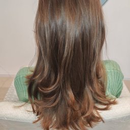 PRACOWNIA FRYZJERSKA ANITA DRATKOWICZ - Brązowe, wycieniowane włosy z refleksami, widok z tyłu. Klientka w zielonym swetrze siedzi na białym fotelu w salonie. Delikatne fale na końcach.
