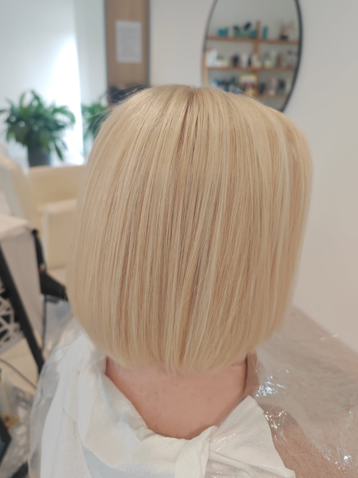 Blond włosy po koloryzacji i strzyżeniu na boba, widok z tyłu, w salonie. Delikatne refleksy i gładka tekstura włosów. Na ramionach peleryna ochronna.
