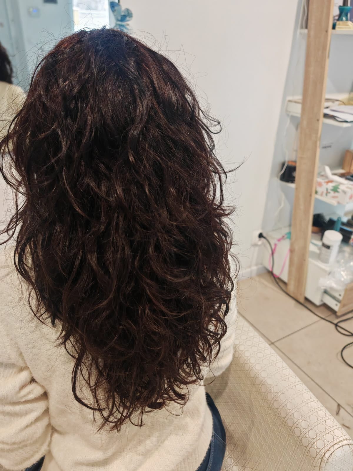 Ciemne, kręcone włosy po zabiegu stylizacji, widoczne od tyłu. W tle jasne wnętrze salonu z półkami i akcesoriami.