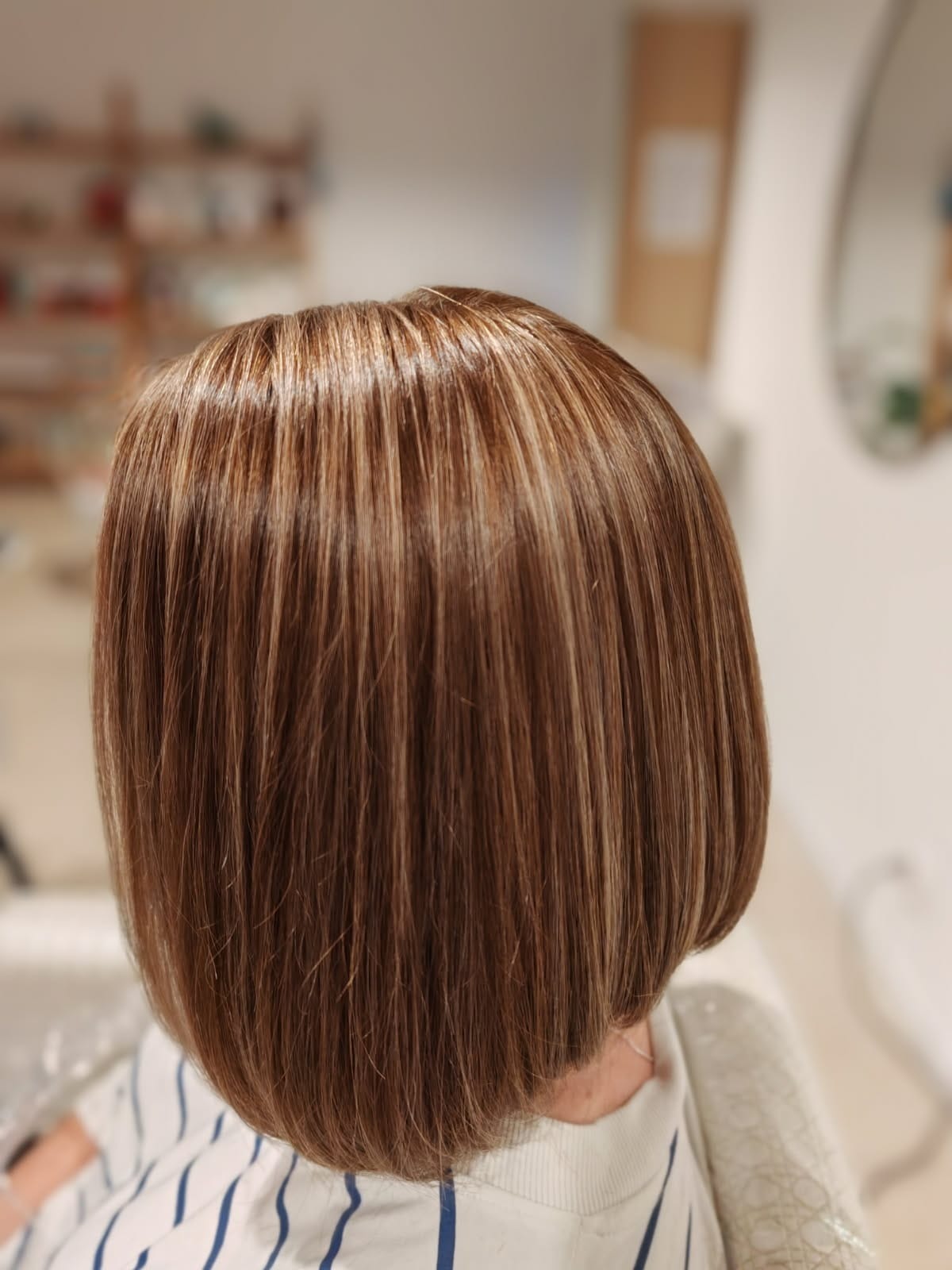 Brązowe włosy z blond pasemkami, krótko obcięte w stylu bob, widoczne od tyłu, z delikatnym połyskiem i naturalnym światłem w tle.