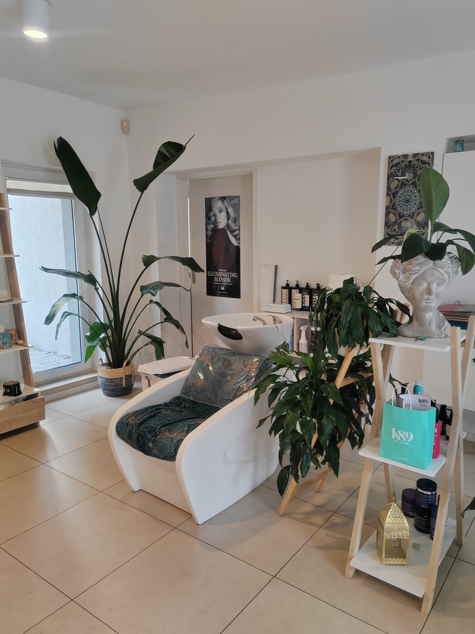 Wnętrze salonu z fotelem do mycia włosów, roślinami doniczkowymi i regałem z kosmetykami. Minimalistyczny wystrój z akcentami zieleni i bieli.