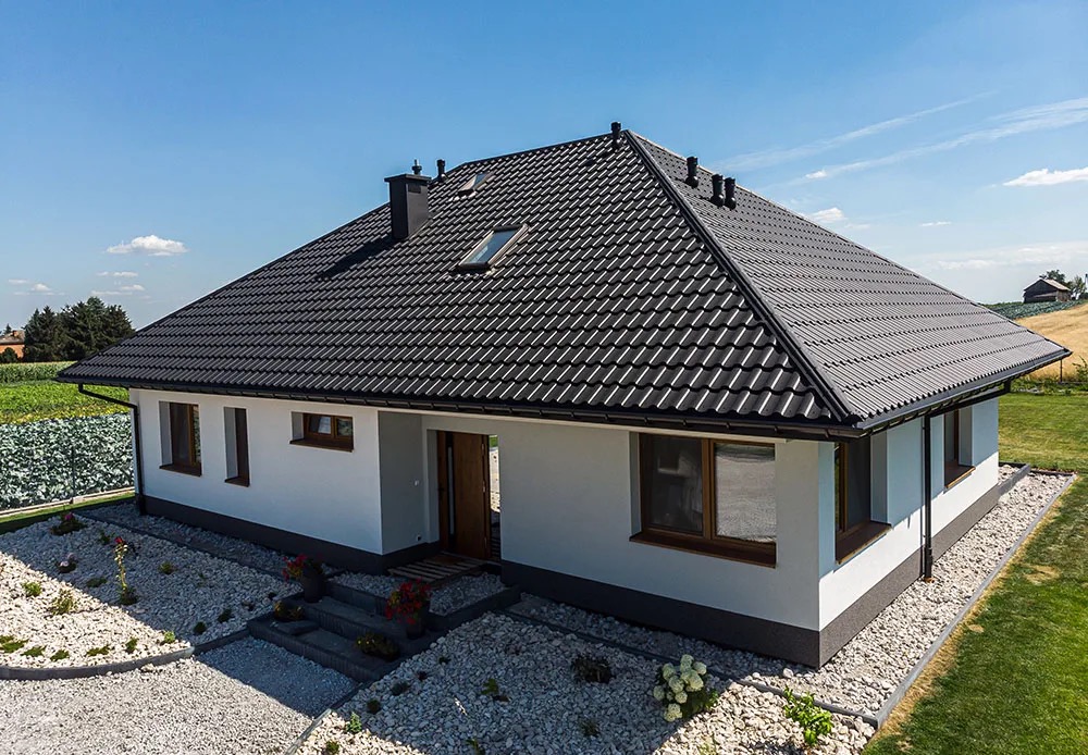 Nowoczesny dom jednorodzinny z ciemną, skośną dachówką, widok z góry. Dach z oknem połaciowym, komin. Biała elewacja, kamienny ogródek i zielone otoczenie.