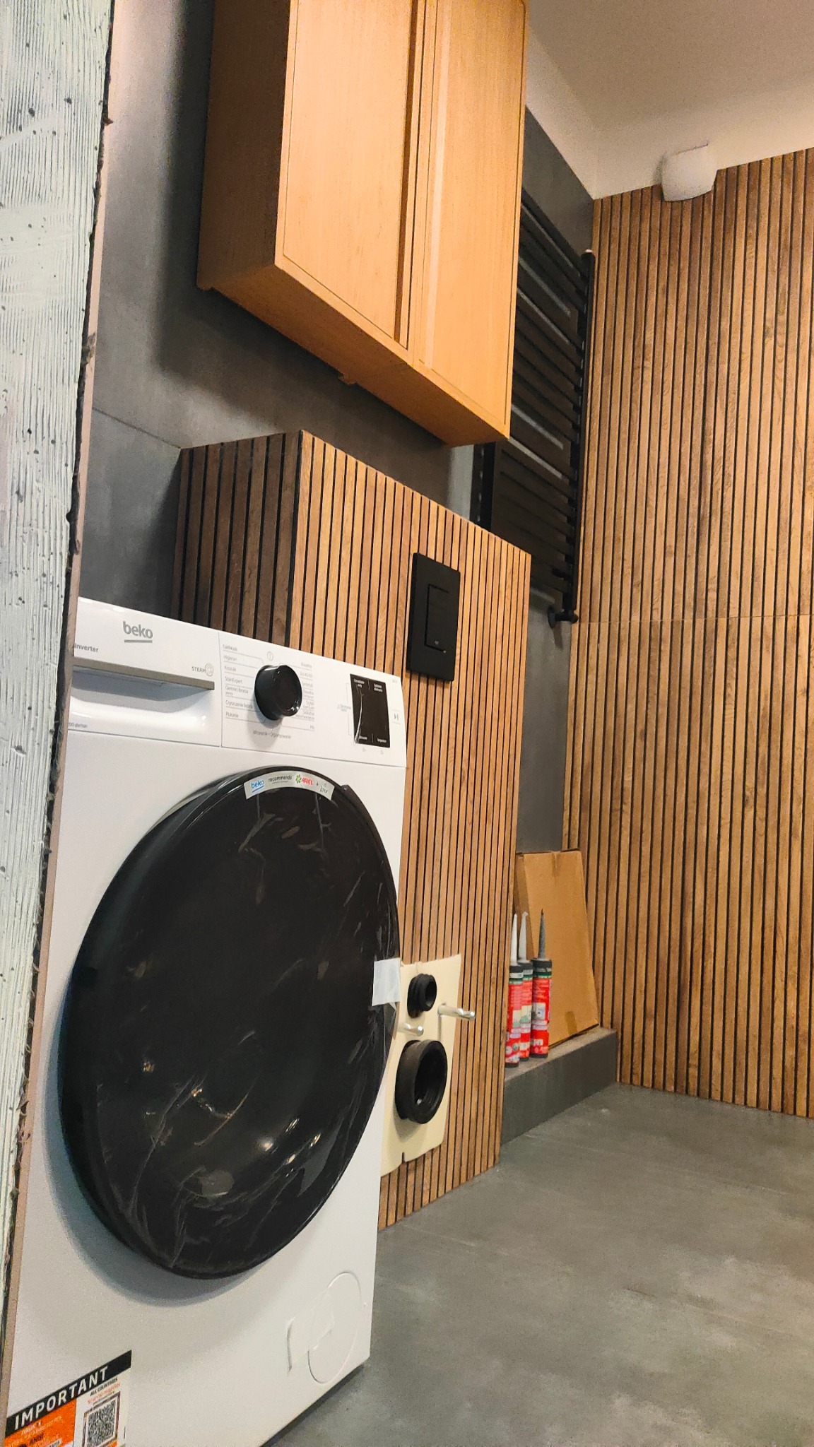 Nowoczesna pralnia z drewnianą boazerią, szafką i grzejnikiem. Biała pralka Beko z czarnymi drzwiami, uszczelniona silikonem. Szare ściany i podłoga.