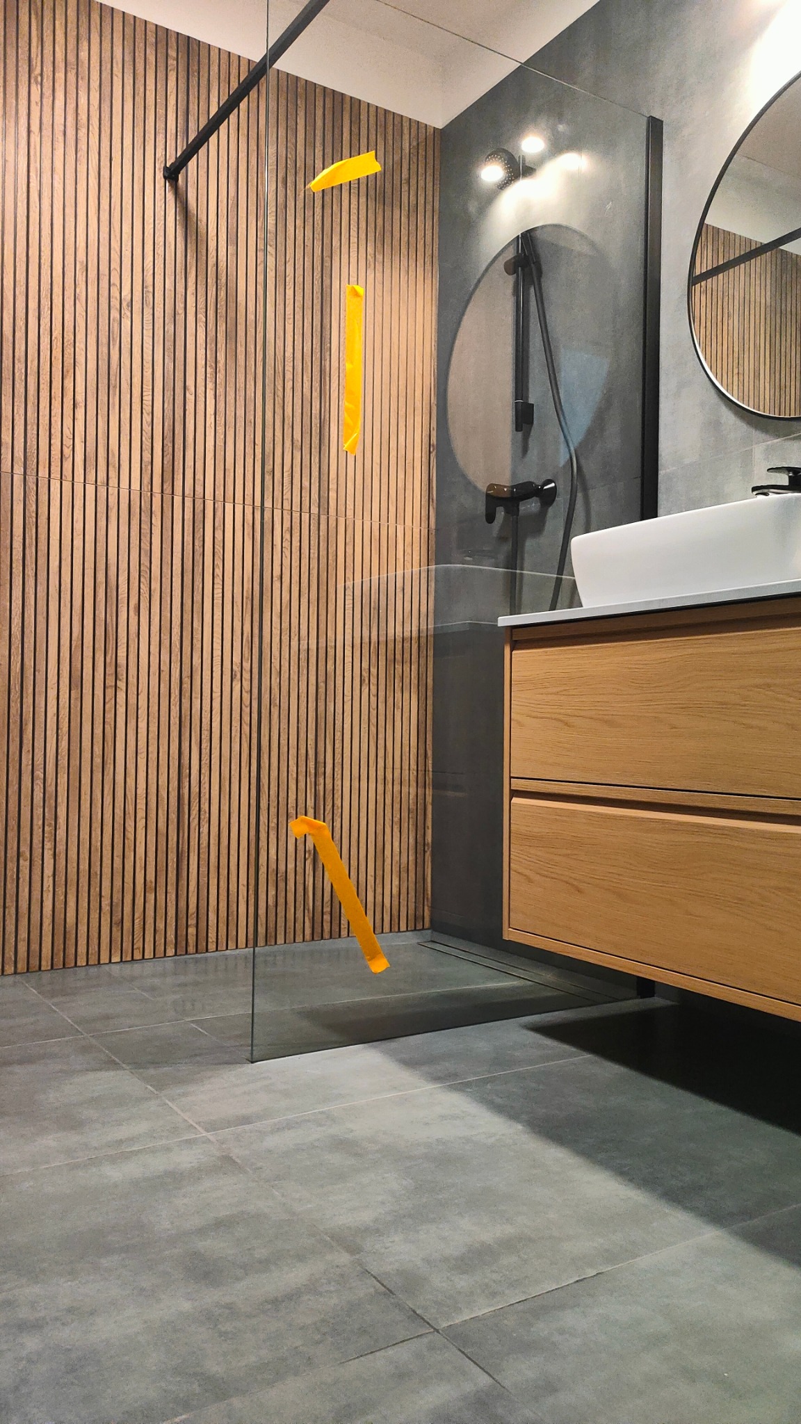 Nowoczesna łazienka z drewnianą ścianą, szklaną kabiną prysznicową i drewnianą szafką pod umywalką. Minimalistyczny design, szare płytki podłogowe.