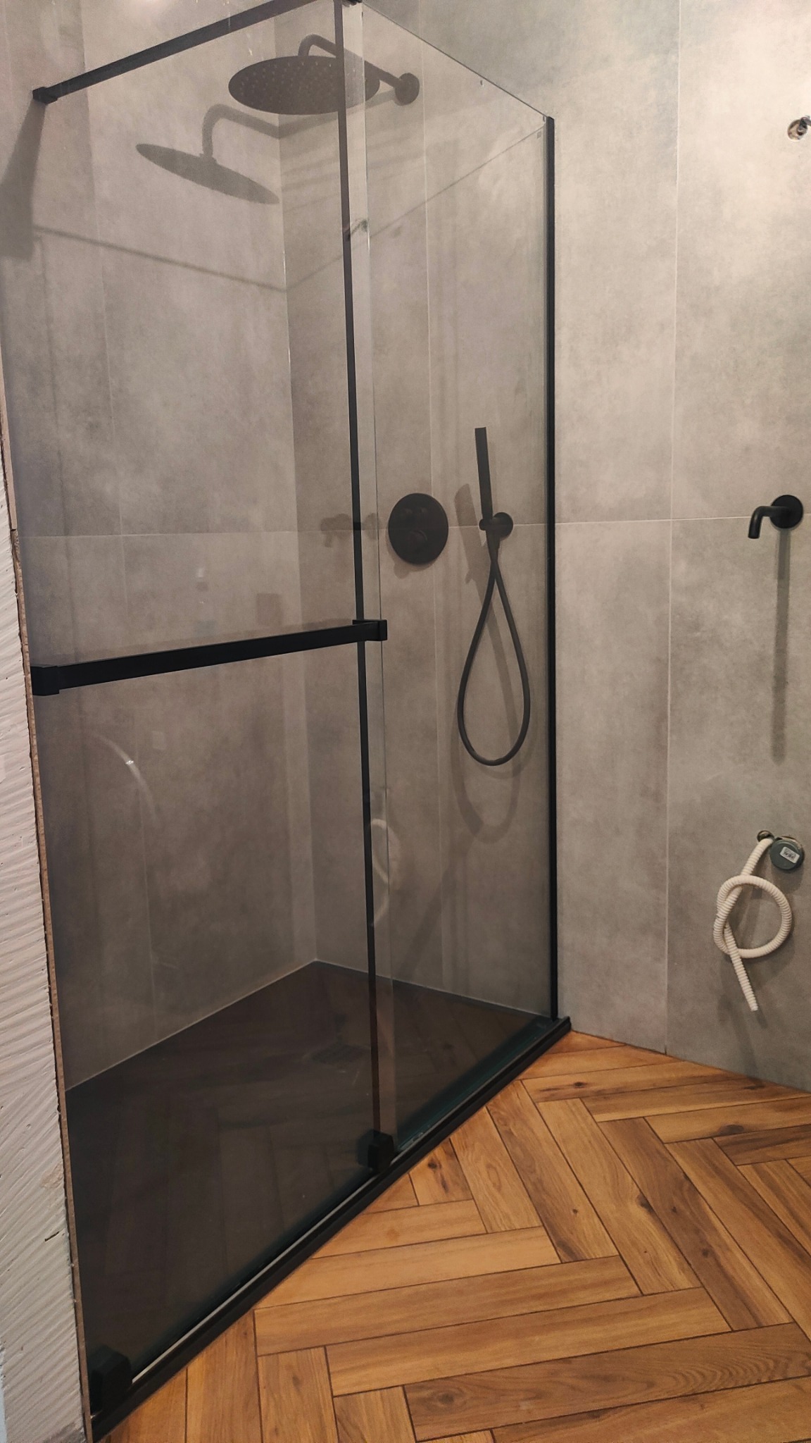 Nowoczesna kabina prysznicowa z czarnymi elementami i deszczownicą, na tle szarych płytek i drewnianej podłogi ułożonej w jodełkę, z widoczną instalacją wodną.
