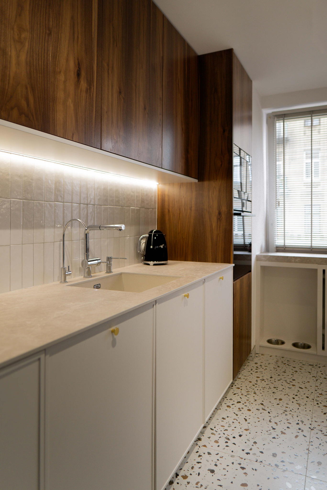 Nowoczesna kuchnia: drewniane szafki, białe fronty, złote gałki, terazzo na podłodze i subtelne oświetlenie LED pod szafkami wiszącymi.