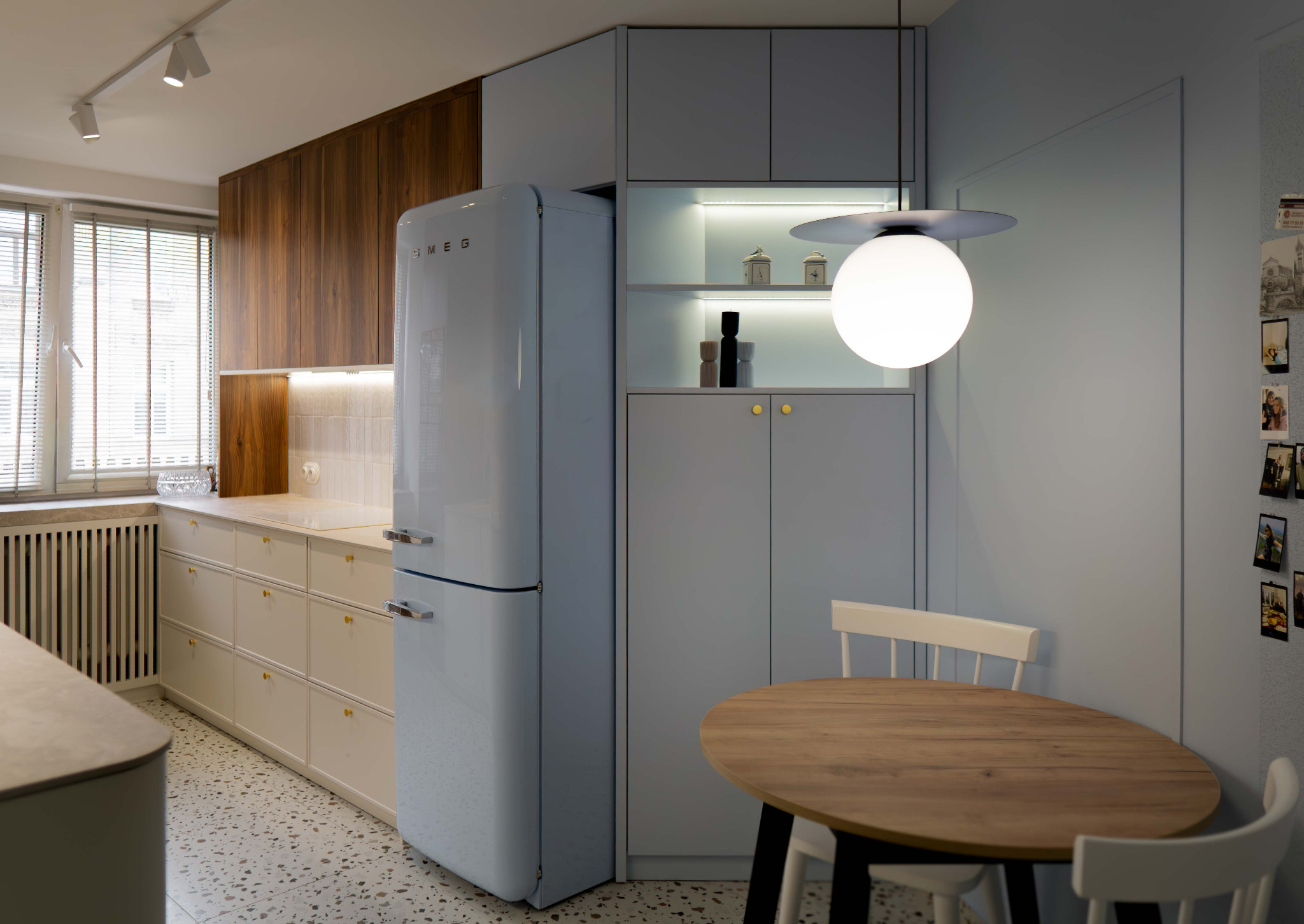 Stylowa kuchnia w błękitach i drewnie: lodówka Smeg, okrągły stół z krzesłami, drewniane szafki i terazzo na podłodze. Nowoczesne oświetlenie.
