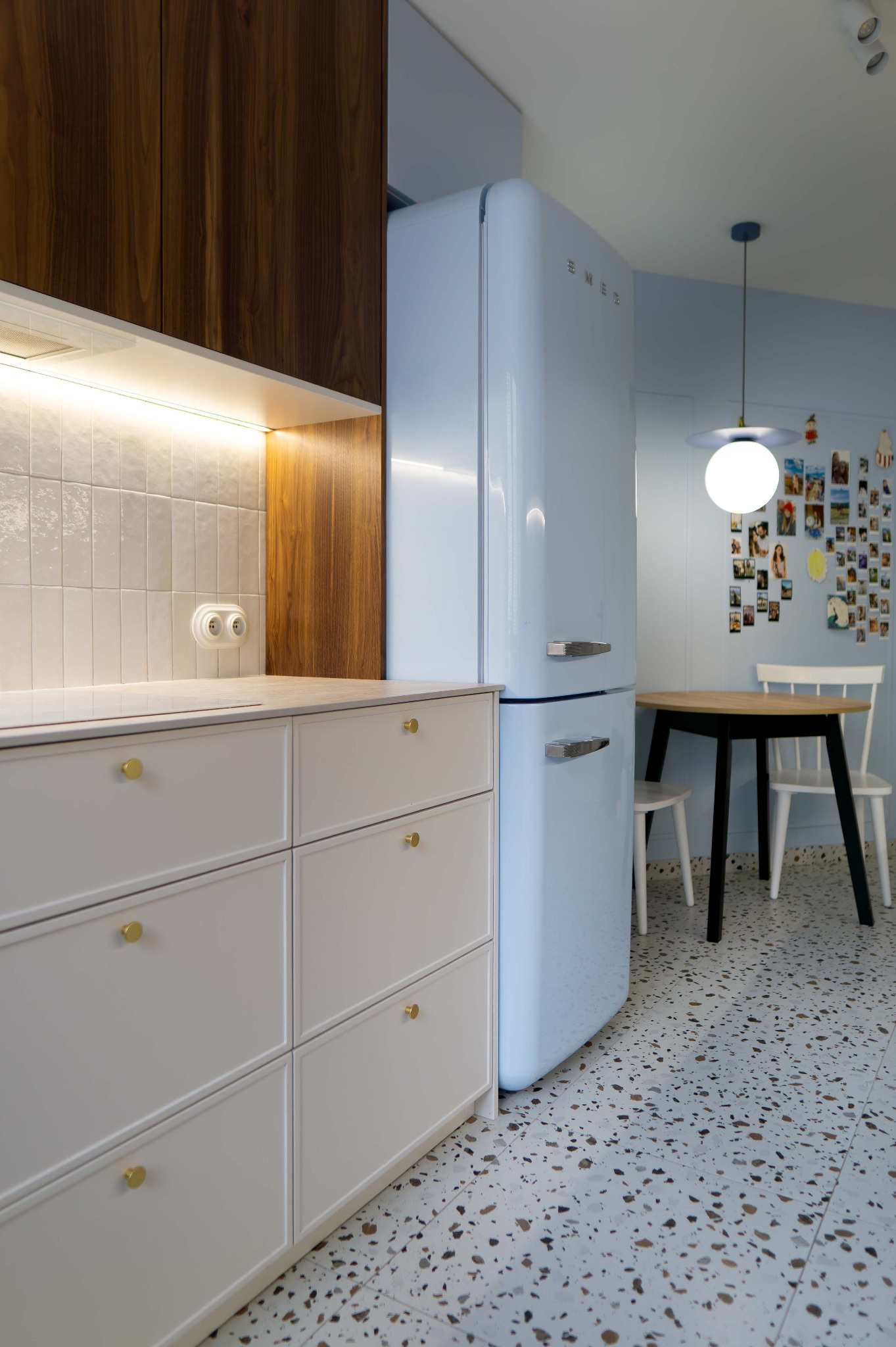 Jasna kuchnia z błękitną lodówką SMEG, białymi szafkami z złotymi uchwytami i mozaikową podłogą. Na ścianie zdjęcia i okrągły stół z krzesłami.