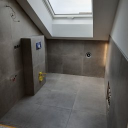 Remigiusz Poczwardowski - Wykończona łazienka z szarymi płytkami, oknem dachowym i elementami instalacji. Widoczne przyłącza wodne i stelaż podtynkowy WC. Minimalistyczny design.