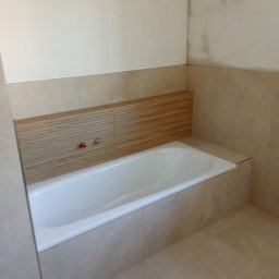 Remigiusz Poczwardowski - Wykończona łazienka z wanną obudowaną beżowymi płytkami, drewniany panel nad wanną, beżowe płytki na ścianach i podłodze. Minimalistyczny design.