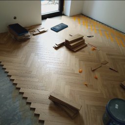 Remigiusz Poczwardowski - Układanie parkietu w jodełkę: widok z góry na częściowo ułożoną podłogę, narzędzia i materiały przygotowane do dalszych prac. Widoczne żółte linie pomocnicze.