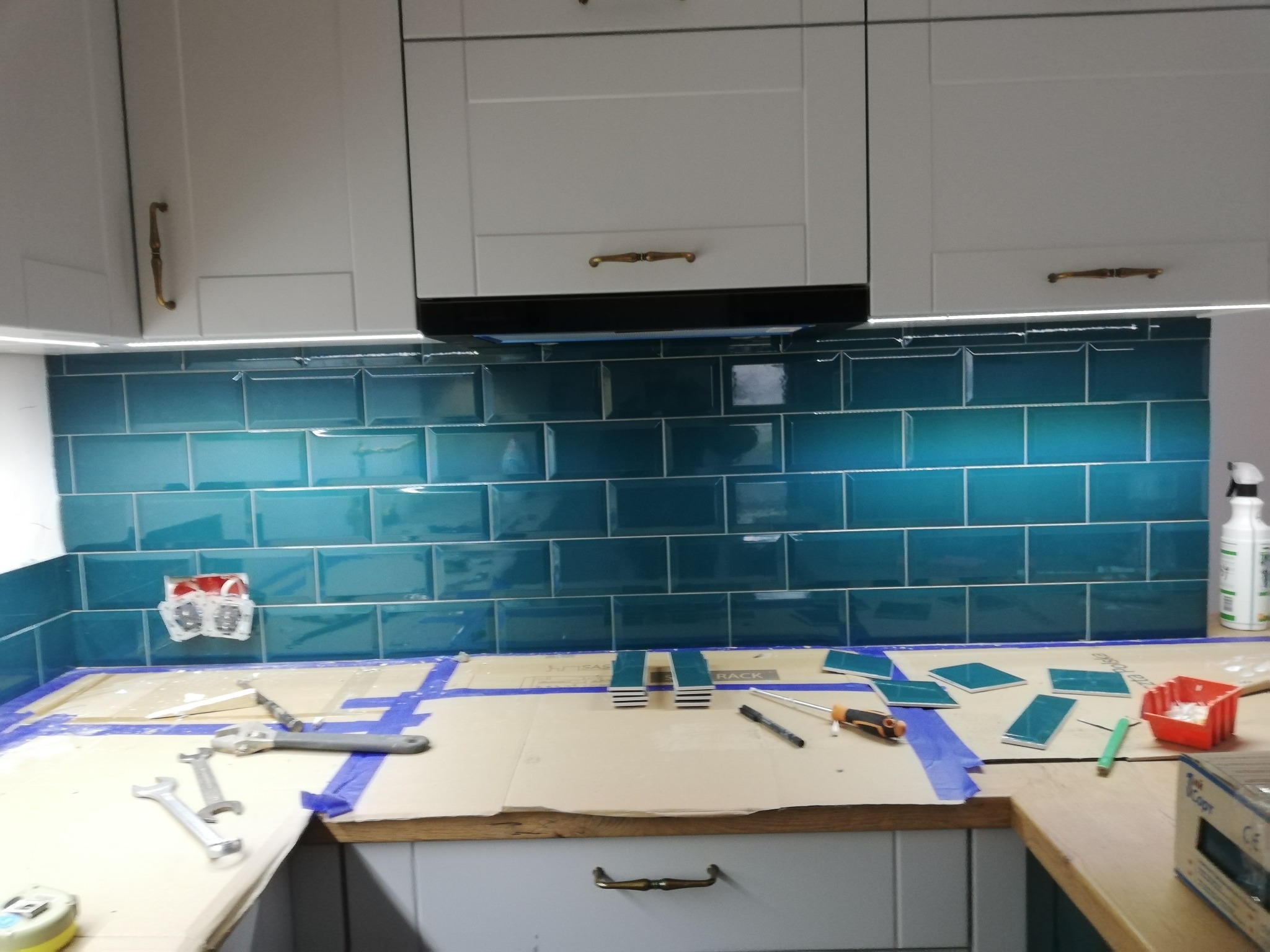Turkusowe płytki ułożone na ścianie kuchni, widoczne narzędzia glazurnicze i zabezpieczone blaty. Etap prac wykończeniowych, oświetlenie LED pod szafkami.