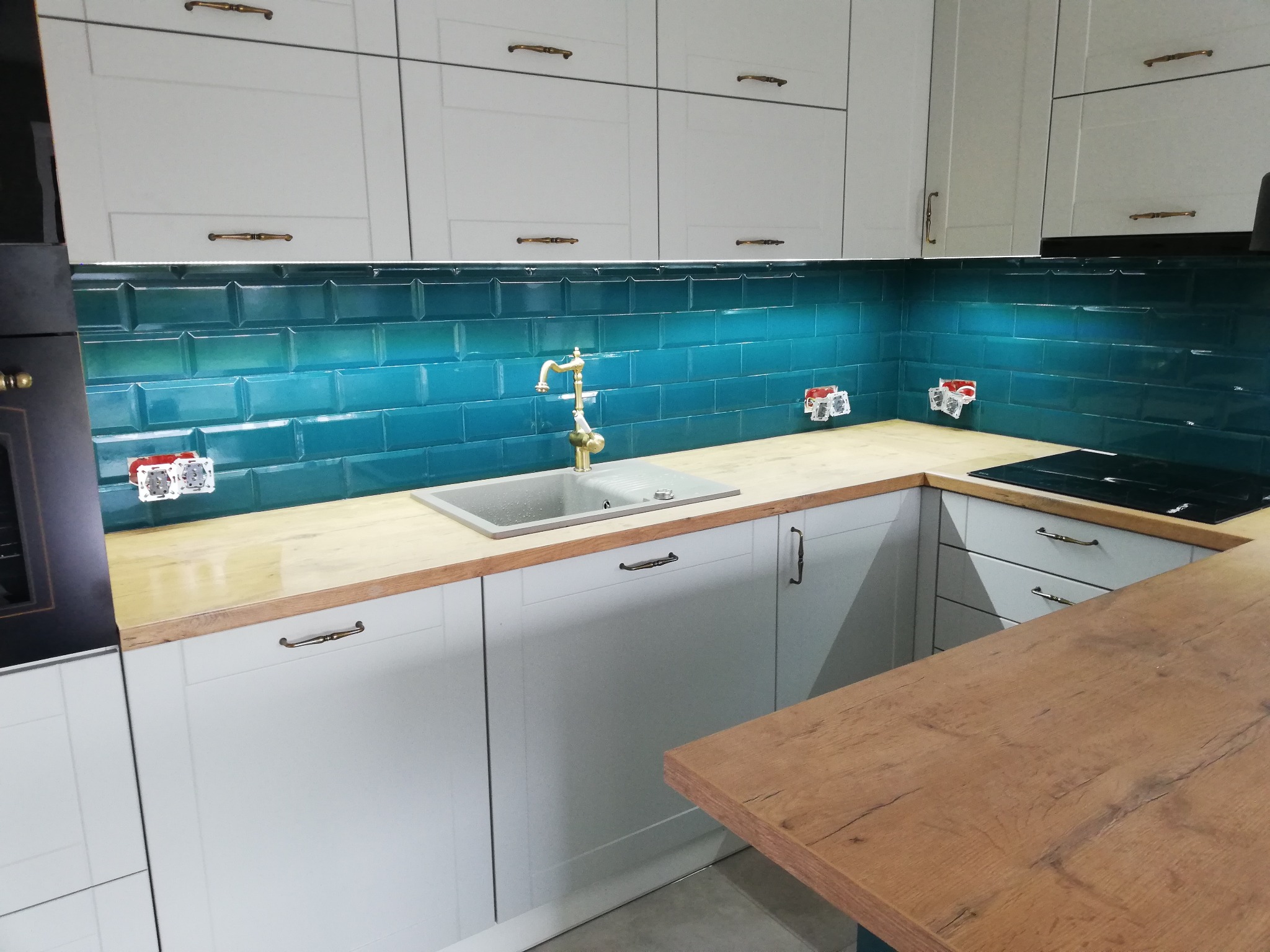Nowoczesna kuchnia z turkusowymi płytkami typu metro, złotą armaturą i białymi szafkami z ozdobnymi uchwytami. Widoczne gniazdka elektryczne nad blatem.