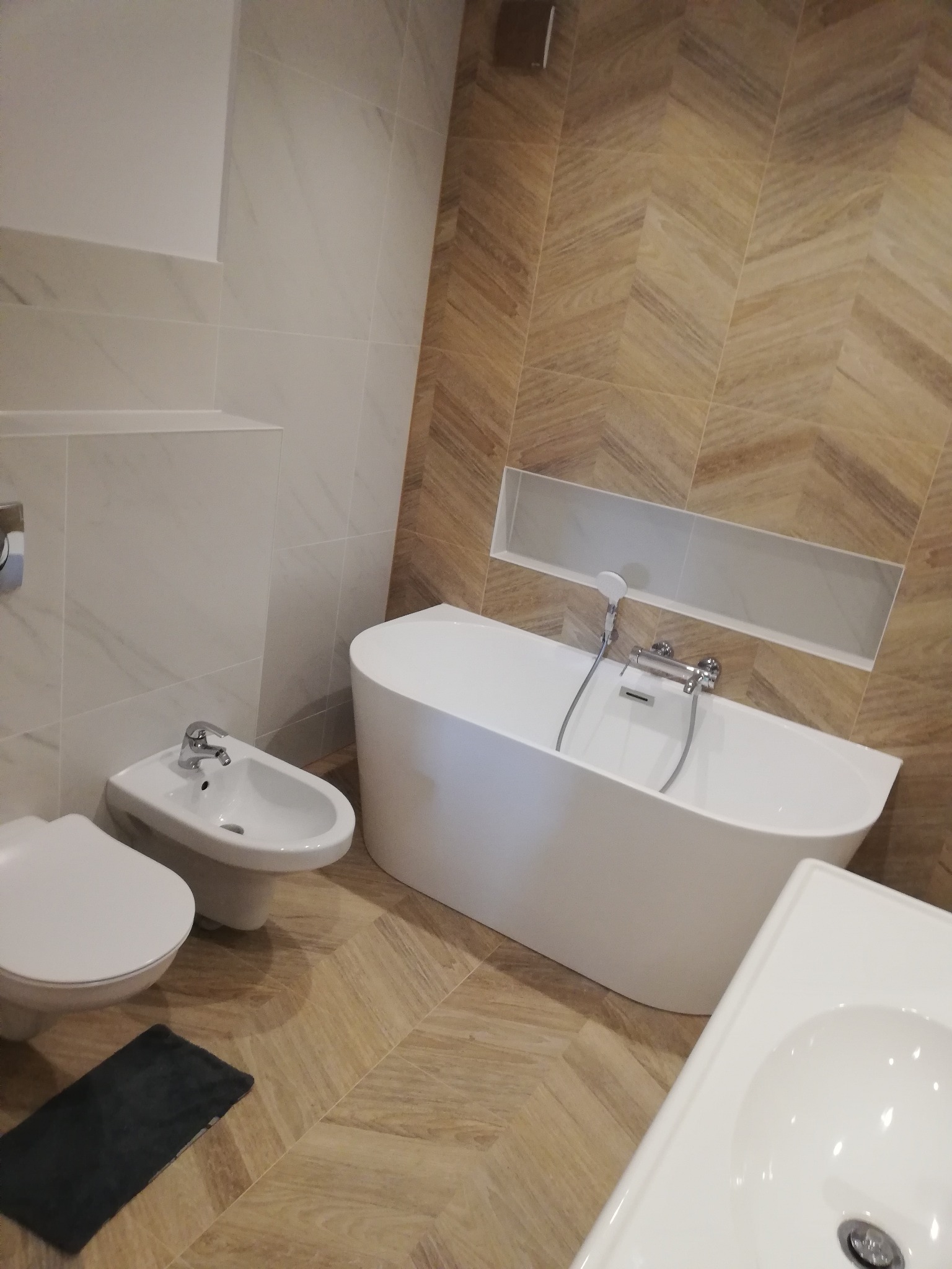 Nowoczesna łazienka z beżowymi płytkami ułożonymi w jodełkę, białą wanną wolnostojącą, bidetem i umywalką. W ścianie wnęka z oświetleniem.