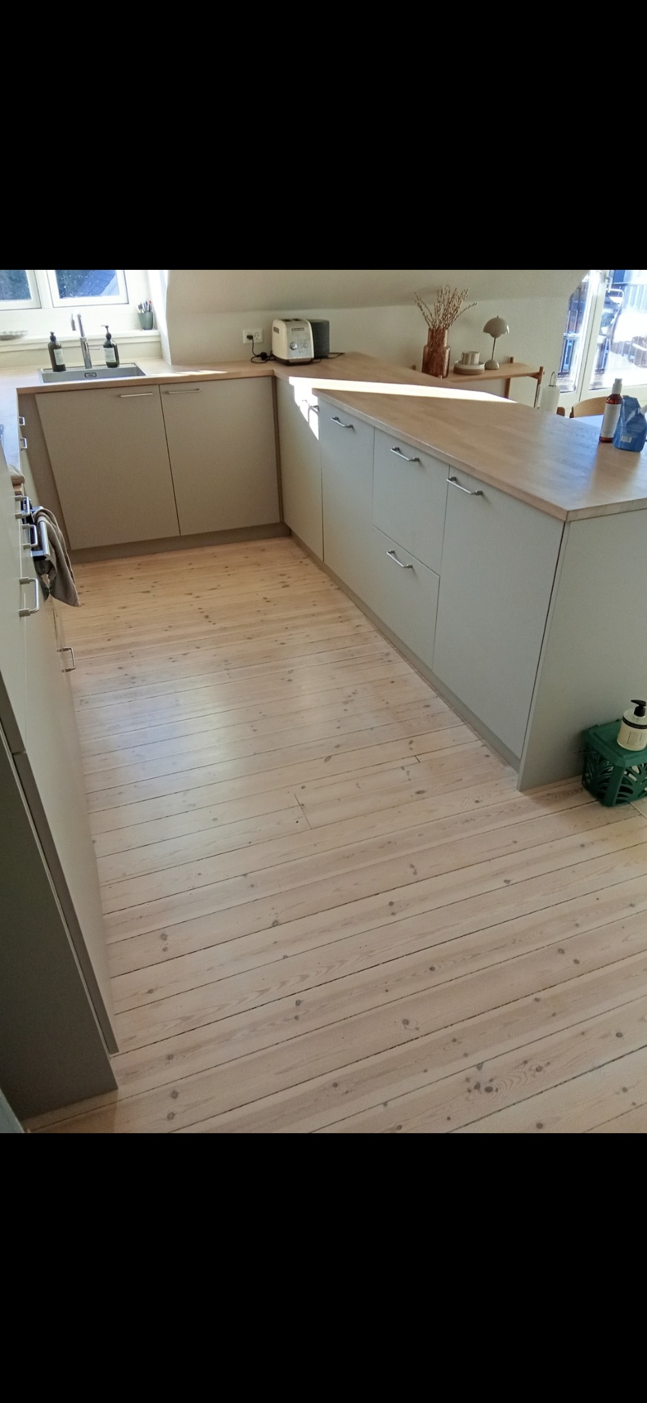 Jasna kuchnia z drewnianą podłogą i szarymi szafkami. Na blacie toster i wazon. Widok na okno z zewnątrz. Minimalistyczny design.