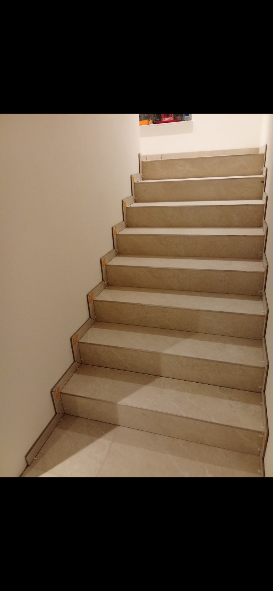 Wykończone schody z beżowych płytek, zabezpieczone taśmą malarską. Widok z dołu, jasne ściany, minimalistyczny styl. Detal wykończenia wnętrza.