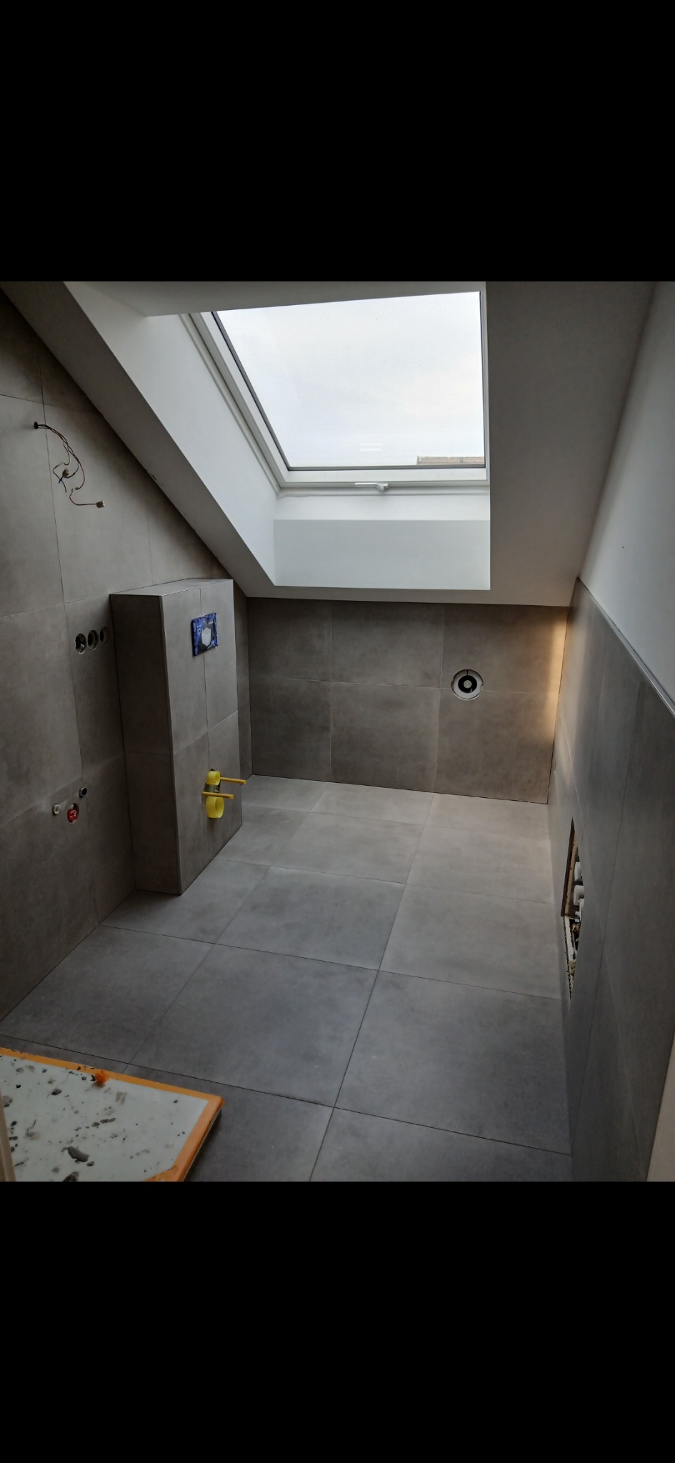 Wykończona łazienka z szarymi płytkami, oknem dachowym i elementami instalacji. Widoczne przyłącza wodne i stelaż podtynkowy WC. Minimalistyczny design.