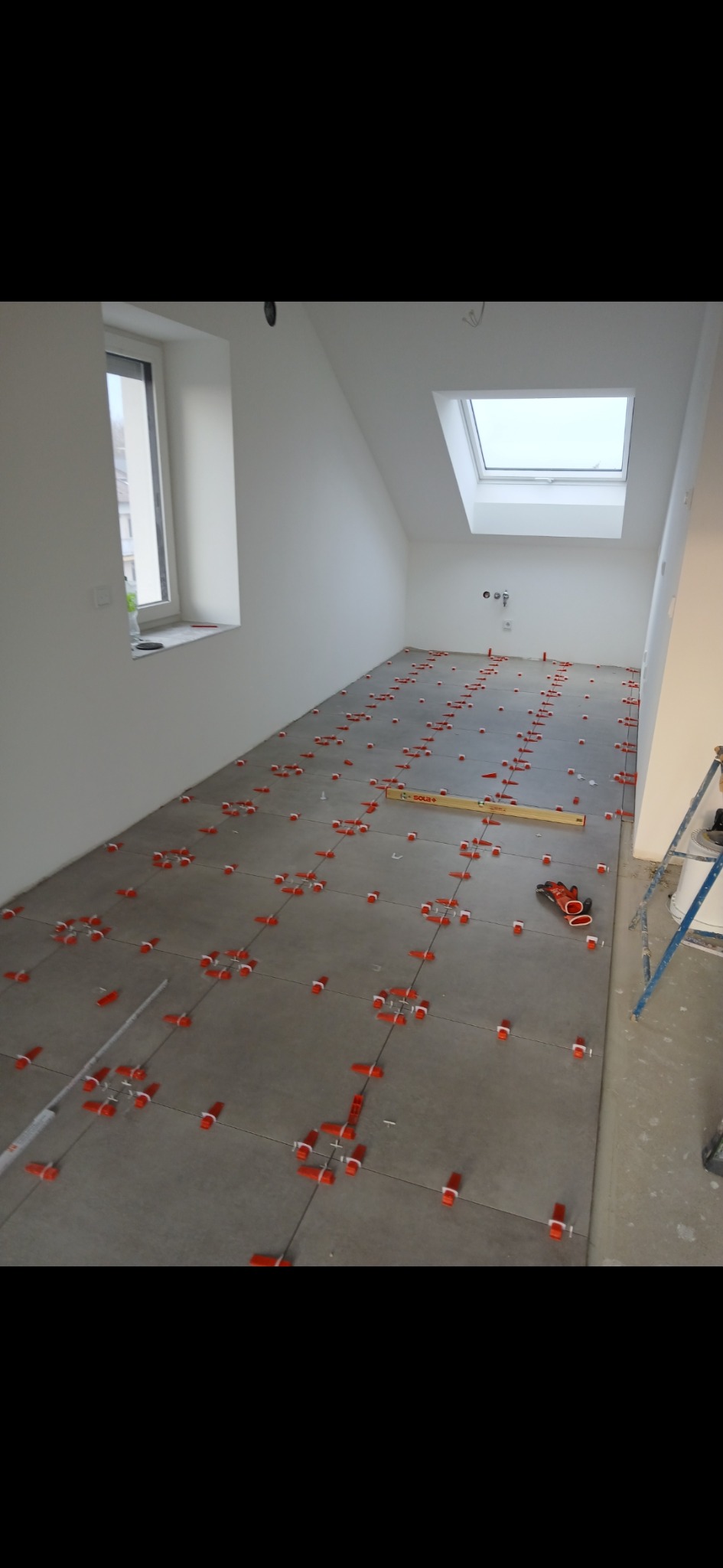 Szare płytki podłogowe w trakcie układania, z czerwonymi krzyżykami dystansowymi, poziomica i okno dachowe w jasnym pomieszczeniu na poddaszu.