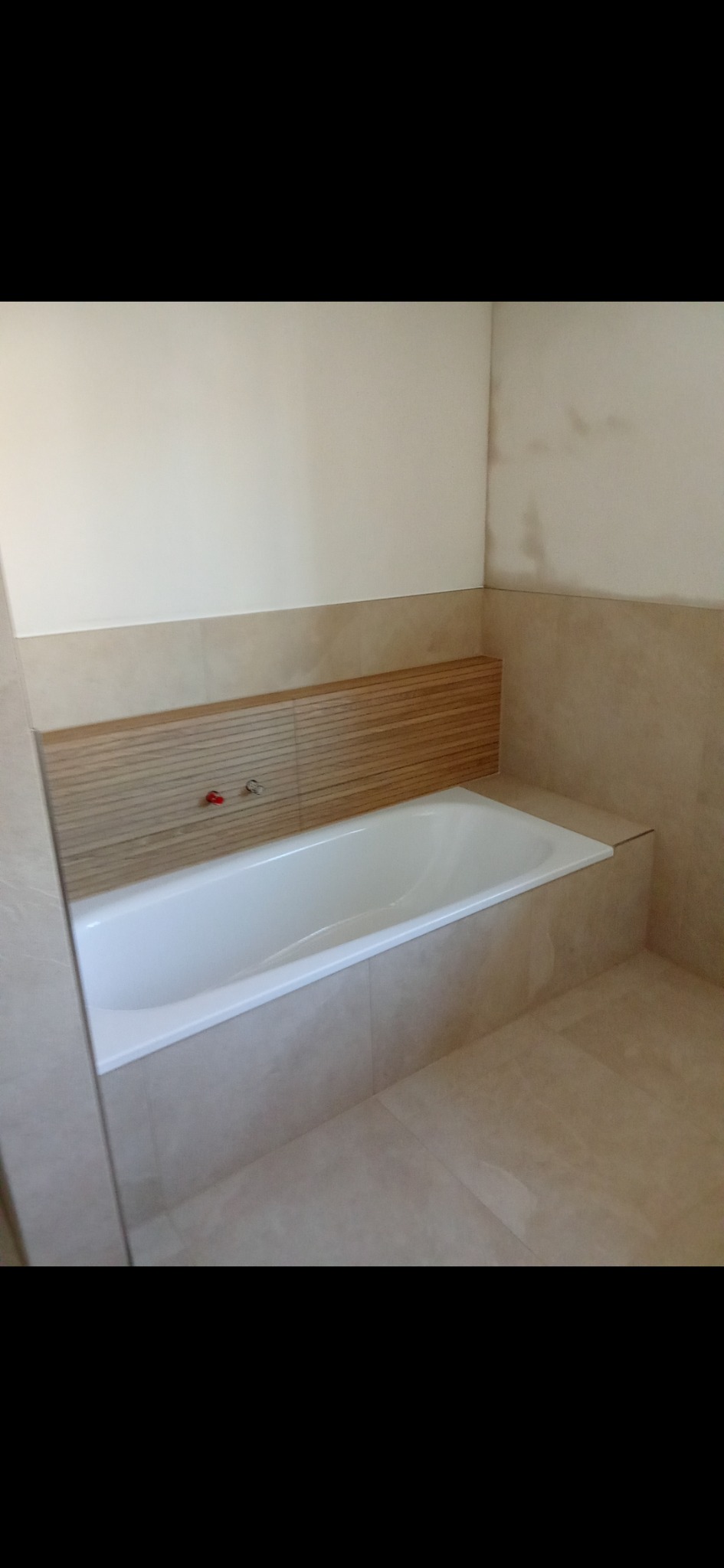 Wykończona łazienka z wanną obudowaną beżowymi płytkami, drewniany panel nad wanną, beżowe płytki na ścianach i podłodze. Minimalistyczny design.