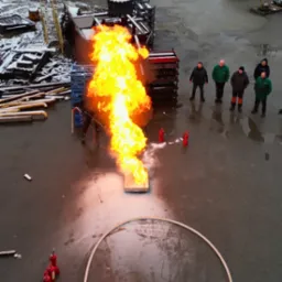 Z góry: Instruktor gasi pożar na poligonie podczas pokazu dla grupy osób. Widoczne gaśnice, płomienie, sprzęt i zimowy krajobraz w tle. Szkolenie ppoż.