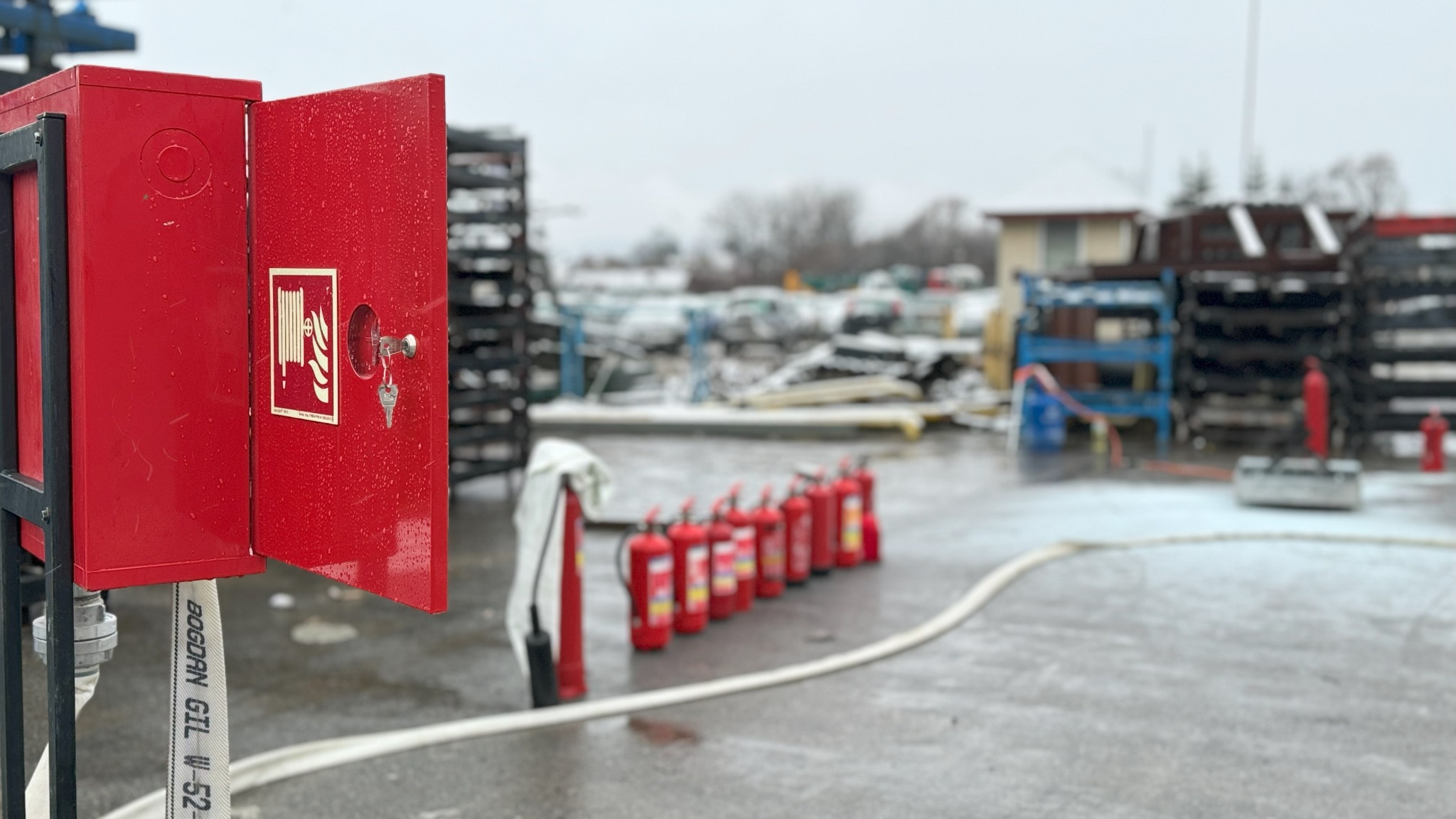 Czerwona szafka hydrantowa z otwartymi drzwiczkami i kluczem, szereg gaśnic i wąż na zewnątrz w zimowych warunkach. Sprzęt ppoż na placu.