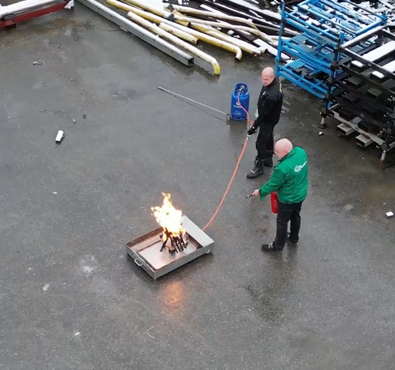 Szkolenie z gaszenia ognia: instruktorzy demonstrują użycie gaśnicy na tacy z płomieniami. Widoczna butla gazowa i palety w tle. Ujęcie z góry.