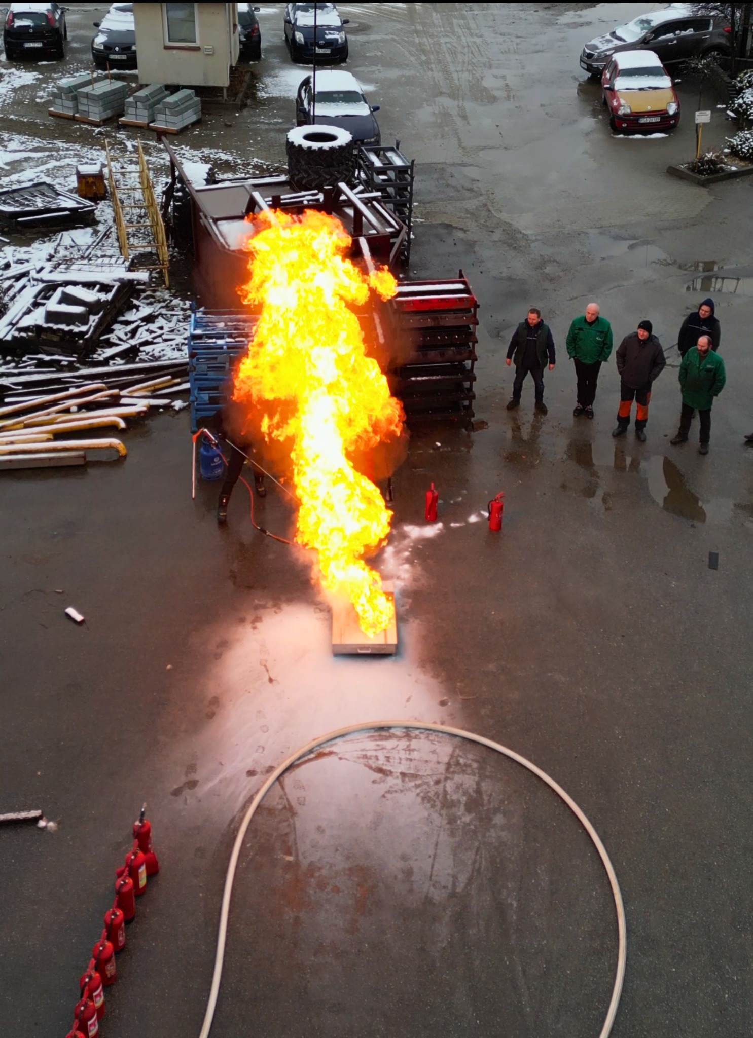 Z góry: Instruktor gasi pożar na poligonie podczas pokazu dla grupy osób. Widoczne gaśnice, płomienie, sprzęt i zimowy krajobraz w tle. Szkolenie ppoż.